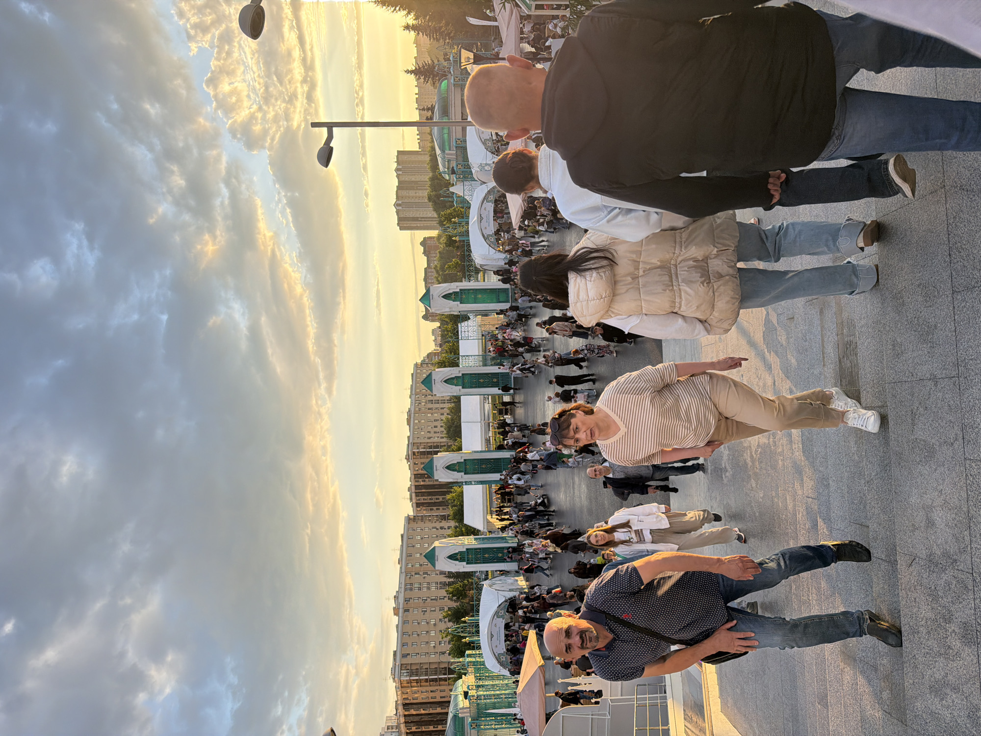 This media file depicts an outdoor event with a large gathering of people. The perspective is from a slightly elevated position, looking down and across a paved area with multiple levels and structures. The sun is low in the sky, casting a warm golden light and creating long shadows, indicating it is either early morning or late afternoon. The sky is filled with dramatic clouds, some illuminated by the setting sun, suggesting fair weather with some cloud cover.
In the foreground, several individuals are visible, including a man in a dark patterned shirt and jeans, and a woman in a striped shirt and tan pants. Other people are seen further back, forming a crowd that stretches into the middle ground and background. There are several white tent-like structures with green ornate entrances, possibly part of a festival or market. Buildings with multiple windows are visible in the distance, hinting at an urban or suburban setting. The overall atmosphere appears to be lively and social, with people casually dressed and engaged in an outdoor event. The image seems to capture a moment of communal gathering under a picturesque sky.