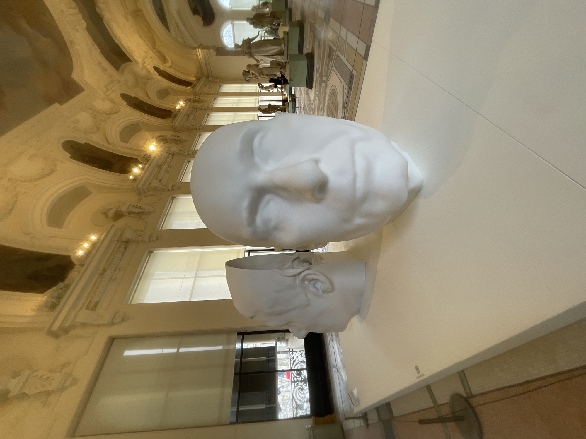 The image displays two minimalist white head sculptures positioned on a long, plain white display surface within an ornate museum hall. The sculpture in the immediate foreground presents a frontal view of a smooth, featureless male head. Behind and to its left, a second white sculpture depicts a side profile of a head, appearing as a segmented cast with a visible ear and a vertical cut revealing its interior.
The setting is the grand central hall of the Musée d'Orsay in Paris, France. The background reveals the expansive gallery space with tall, rectangular windows lining the far wall, each fitted with light-colored roller blinds. Above the windows, the architecture features elaborate white stucco moldings, cornices, and decorative arches that extend into a high, barrel-vaulted ceiling. Sections of the ceiling are adorned with painted murals in warm tones, and multiple recessed spotlights provide interior illumination. The floor beyond the display table features a multi-colored geometric mosaic pattern. Numerous classical and neo-classical figurative sculptures, typically in white or light-colored stone, are visible on green or dark pedestals throughout the background gallery, arranged along the patterned floor. No individuals are prominently depicted or engaging with the subjects in the immediate foreground.