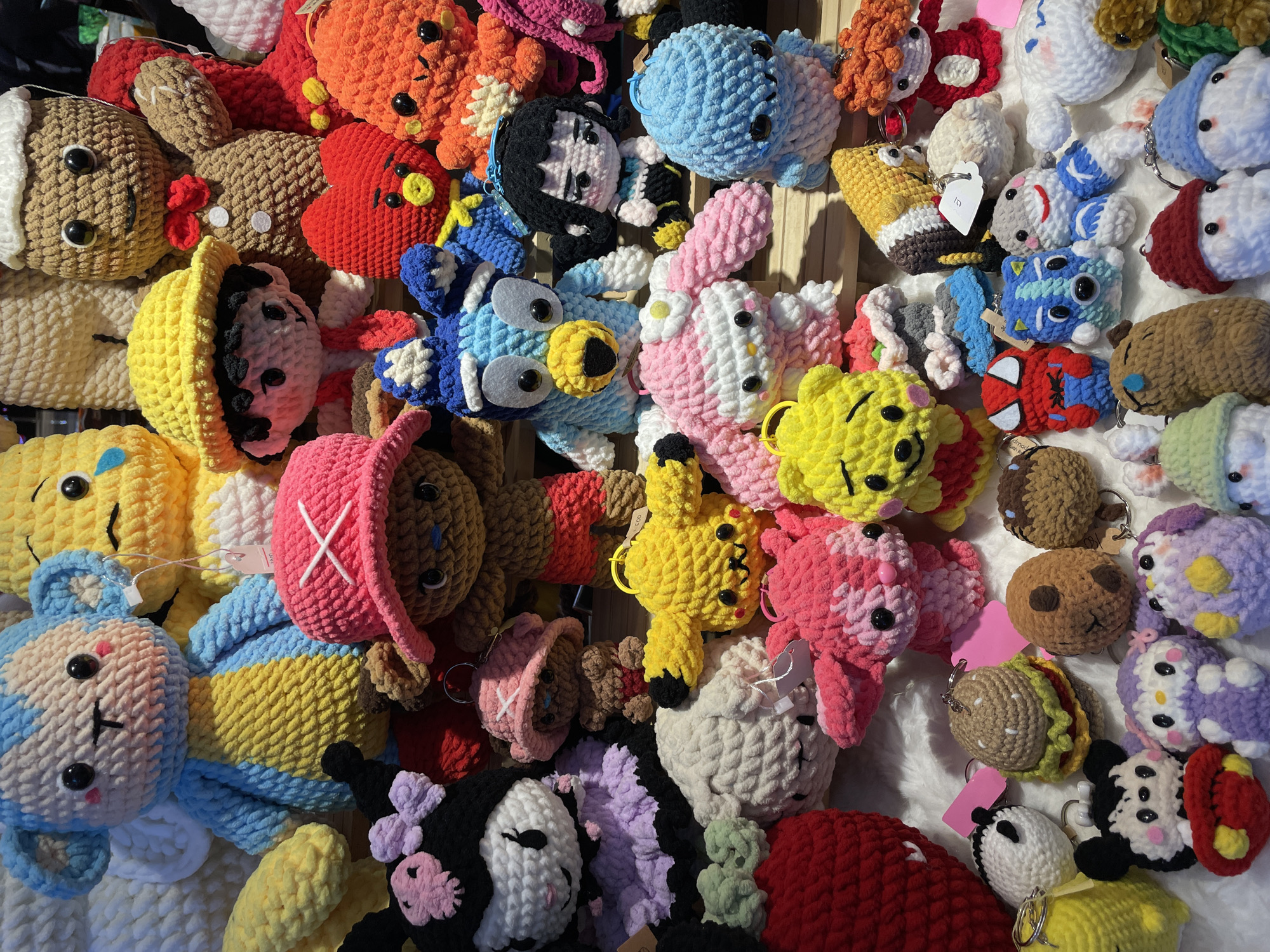 This full overhead shot displays a dense, colorful assortment of handmade crocheted plush keychains and small toys. The items are tightly packed, covering the entire frame, and appear to be arranged on a light-colored, possibly white or cream, surface which might be a fabric-covered table or shelf.
The subjects include a wide variety of cartoon characters, animals, and food items, all rendered in chunky, textured yarn. Notable characters identifiable are Mickey Mouse, Hello Kitty, Spiderman, Bluey, Pikachu, Winnie the Pooh, My Melody, Kuromi, Tony Tony Chopper from One Piece, and several characters from BT21 such as Koya (blue koala) and RJ (white alpaca). Other designs include a gingerbread man, various bunnies, bears, birds, and an item resembling a burger. Many of these items feature attached metal keyrings, indicating their function as keychains or bag charms.
The items are presented for viewing, suggesting a retail setting such as a craft market, stall, or shop. The bright and diverse color palette, including vibrant reds, oranges, yellows, blues, pinks, purples, and browns, contributes to a cheerful and playful atmosphere.
The lighting is bright and even, casting subtle shadows that indicate an overhead light source. Some small white and pink tags are visible attached to a few items, likely indicating prices or branding, though no specific text is clearly legible beyond what might be a number "150" or "100" on one white tag. The overall impression is one of abundance and artisanal craftsmanship.