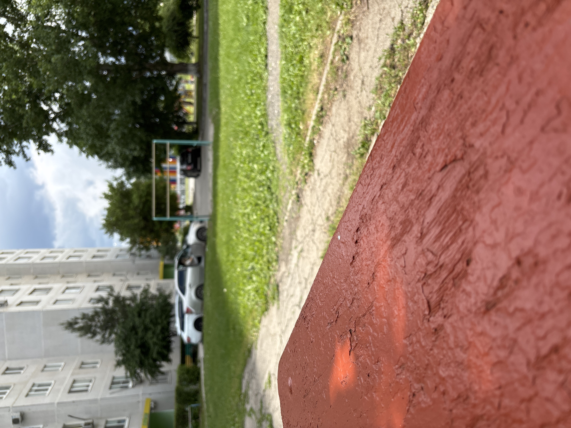 The image shows a residential scene, likely in Lipovka, Russia, given the context. A red painted object, possibly a bench or low wall, dominates the foreground. Beyond, a green grassy area and a walkway lead to a multi-story building with white walls and numerous windows. Parked cars are visible along the walkway. A playground structure can be seen in the distance, partially obscured by trees. The sky is cloudy. The image is taken from a low angle and is slightly rotated.