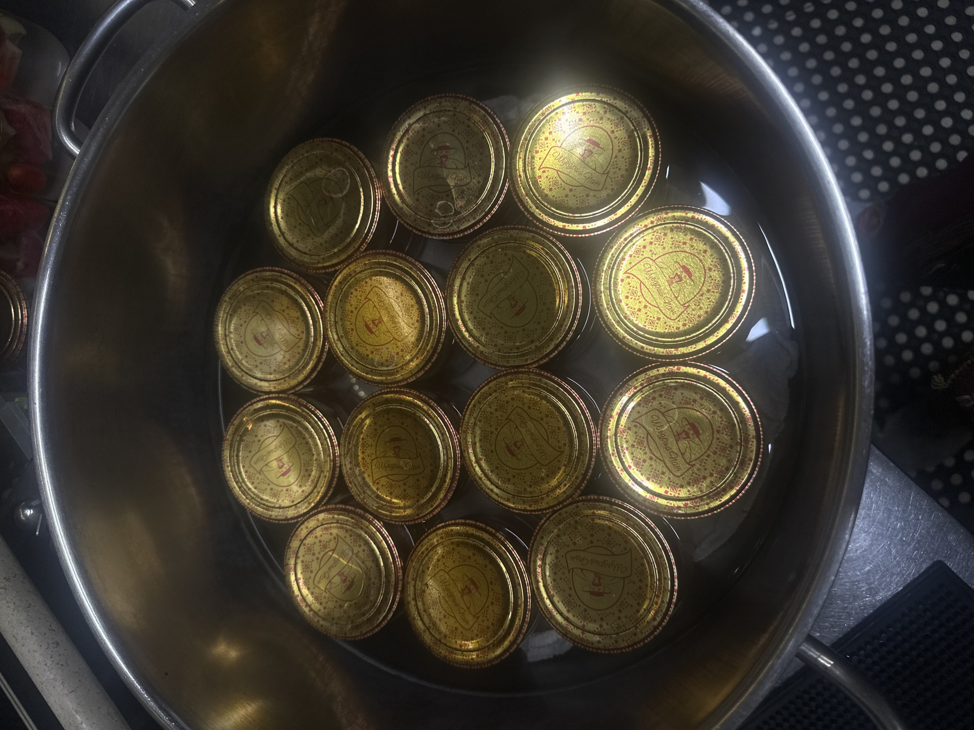 A high-angle shot shows a metal pot filled with water and multiple canning jars with gold lids. The jars are submerged in the water, with the lids facing upwards. The lids are decorated with a red pattern and feature a logo that appears to read "Mrs. Wages' Choice." The pot is sitting on a countertop with a black and white polka-dot surface. The background is blurred, but appears to be a kitchen setting. The image does not contain any information about the city of Ternopil, Ukraine, or any specific location details.