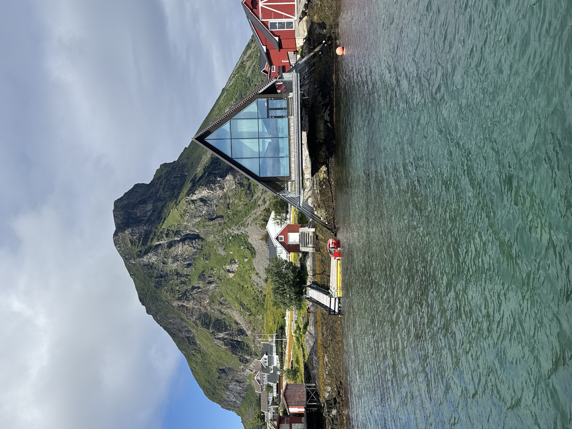 The image shows a scenic view of the Lovund mountain in Lovund, Norway, with a body of water in the foreground and several buildings nestled along the coastline. A prominent A-frame building with a large glass facade stands on stilts over the water, reflecting the sky and surrounding landscape. Adjacent to it is a traditional red Norwegian Rorbu, a fisherman's cabin. Several other white houses are visible further up the hillside, suggesting a small village. The water is a clear, greenish-blue, with small waves. A yellow boat is docked near a wooden pier, and a red motorboat is tied beside it. The mountain is covered in lush green vegetation, with rocky outcrops and a dramatic, imposing peak. The sky is partly cloudy, with patches of blue visible. The overall impression is one of a tranquil, picturesque coastal setting.