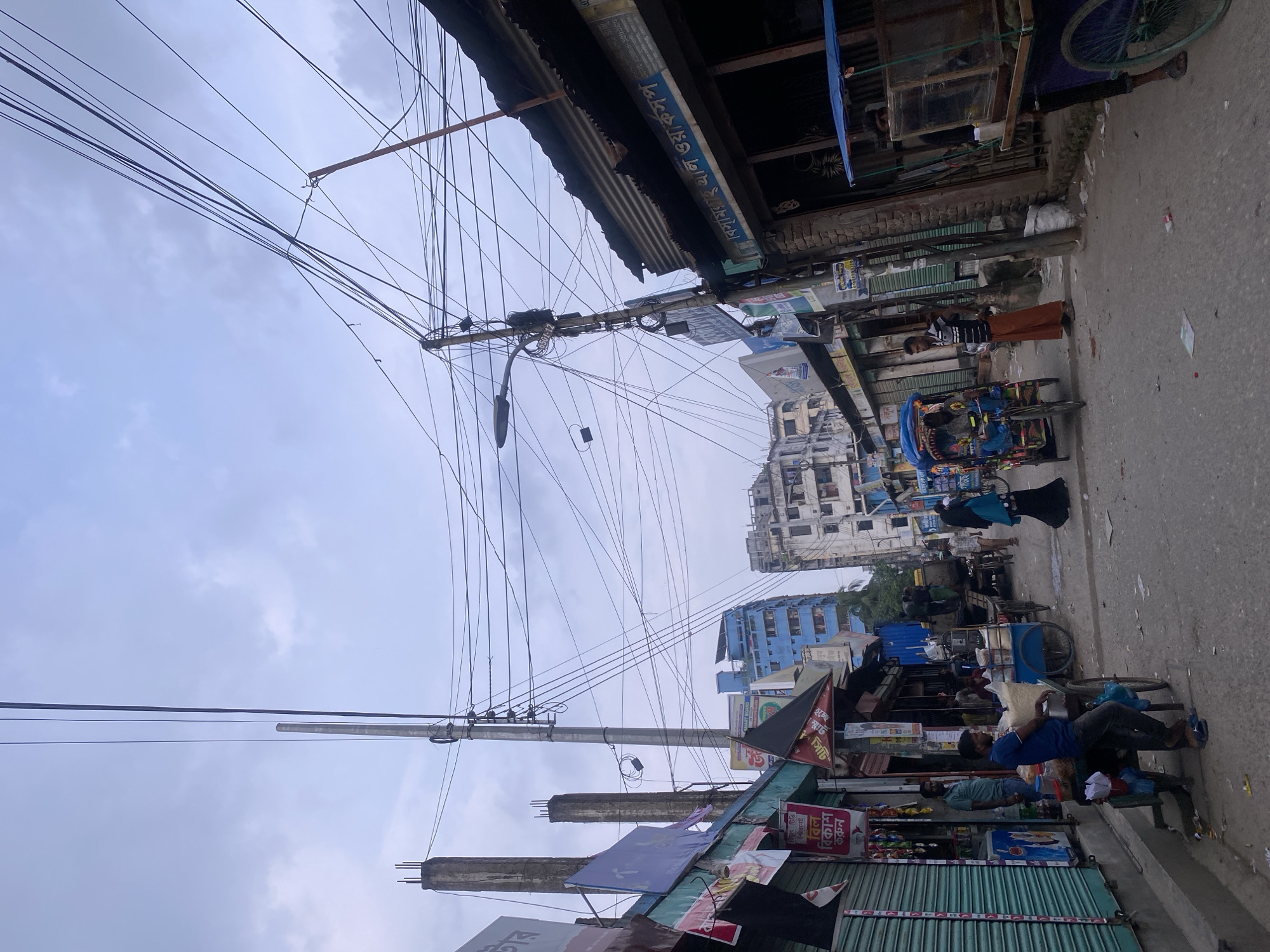 This image captures a street scene in Dhaka, Bangladesh, likely during the daytime under an overcast sky. The street is lined with commercial establishments and residential buildings, with a dense network of overhead electrical wires dominating the sky.
On the left side of the street, various shops are visible, some with their shutters down. Banners and signs, some in Bengali script, are displayed above the shops. A man in a blue shirt and dark pants stands near a cart filled with food items, looking towards the right.
The right side of the street features more shops, one with a corrugated metal roof and a sign that appears to be in Bengali script. A person in a striped shirt and orange pants stands near the entrance of a shop. Further down the street, a rickshaw with a blue canopy is parked. Several people are walking along the street, including a woman in a blue headscarf and black abaya, and another person in a similar outfit further down. A cart with a blue canopy, possibly a food vendor, is also visible.
In the background, multi-story buildings, some appearing modern and blue, stand alongside older, more dilapidated structures. The street is paved and appears to be a busy thoroughfare, with some debris scattered on the ground. The overall impression is of a vibrant, urban environment characteristic of Dhaka.