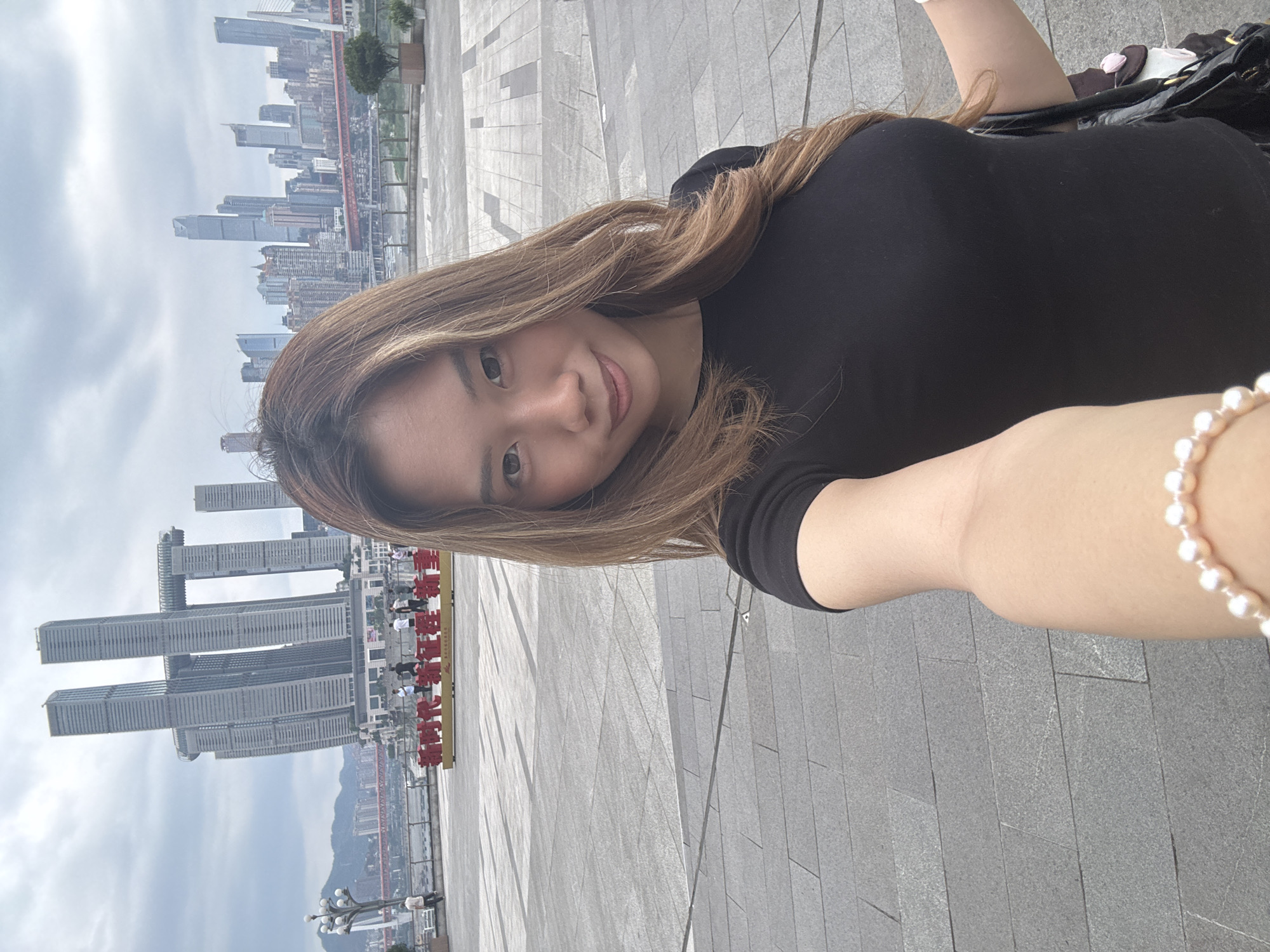 A woman is taking a selfie in Nan'an District, Chongqing, China, with the Raffles City Chongqing complex visible in the background. The woman is wearing a black shirt and a pearl bracelet, and has light brown hair with blonde highlights. She is smiling at the camera. The background shows the urban landscape with gray pavement and numerous high-rise buildings. A red sign with Chinese characters is also visible in the background.