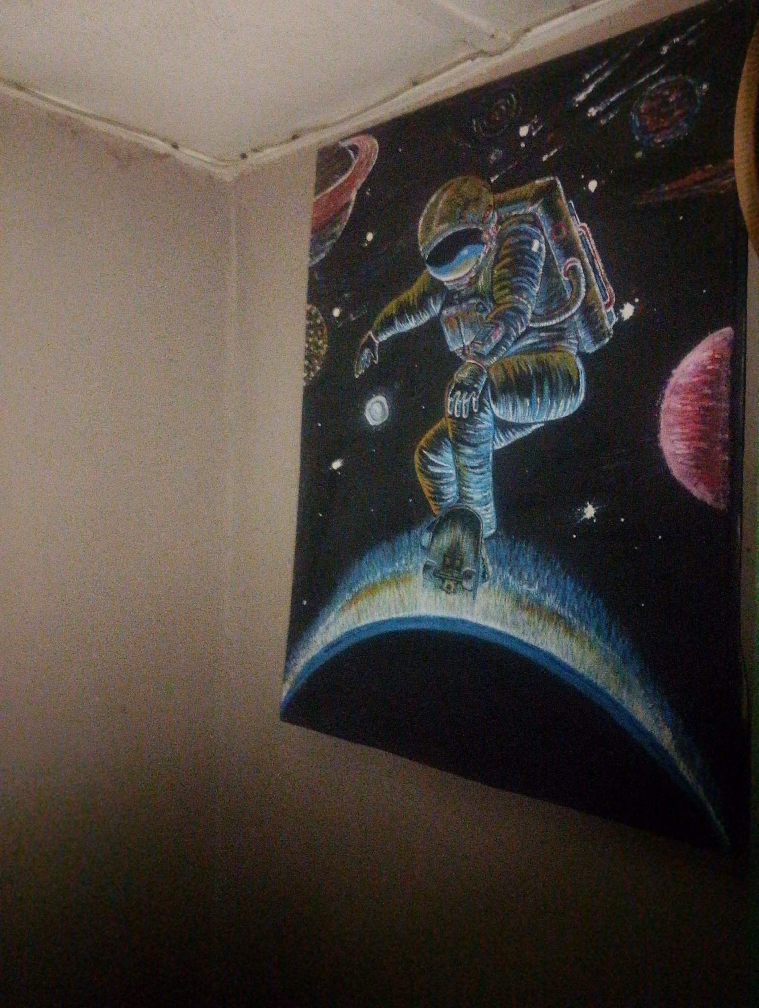 A framed artwork of an astronaut skateboarding in space is mounted on a textured wall in a corner of a room. The artwork depicts a full-body view of an astronaut in a detailed suit, with a helmet that reflects a colorful nebula. The astronaut is posed as if mid-air, with one leg extended, seemingly balancing on a curved, glowing surface that represents a planet's atmosphere or a celestial body. The background is a dark expanse of space, dotted with stars, nebulae, and two planets, one with rings and another a reddish sphere. The artwork itself has a slightly grainy texture, possibly due to the lighting or the medium used. The corner of the room where the artwork is hung shows a junction of two walls and a ceiling, with a white molding running along the top. The walls have a light, textured finish, and the lighting in the room casts a soft, diffused glow on the scene. There are no people visible in the immediate vicinity of the artwork, and the overall impression is that of a decorative piece in a private space, possibly a bedroom or living area. The city of Jos, Nigeria, is not discernible from the visual information presented in the image.