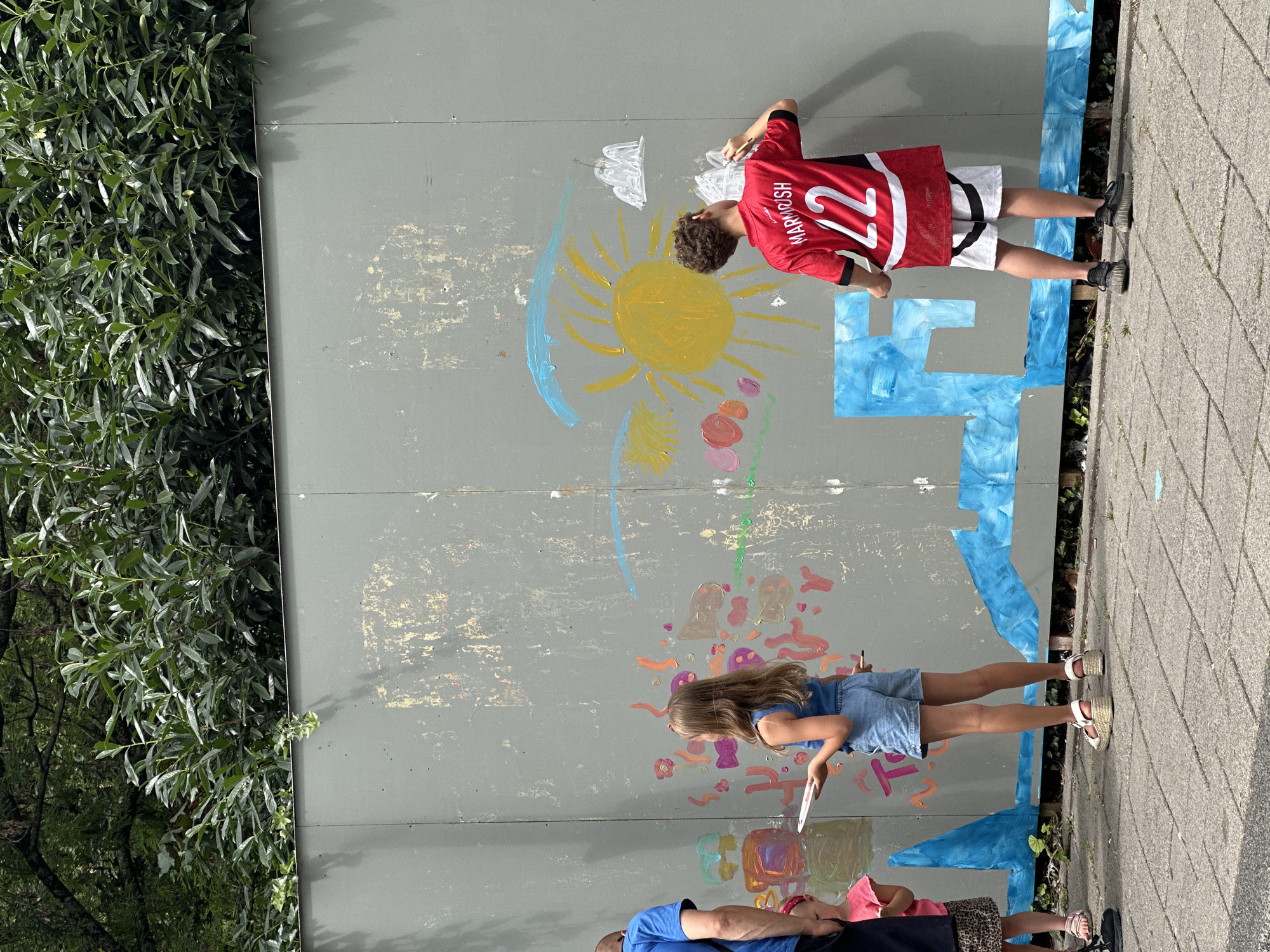 In Amsterdam, Netherlands, a group of children are painting on a large, gray, temporary wall, possibly a construction barrier. A boy in a red jersey with the number "22" is painting a sun with yellow acrylic paint. To his left, a young girl in a blue tank top and denim shorts is holding a plate and a paintbrush, looking at the wall. Further to the left, another child with a pink headband is partially visible. The wall is adorned with various colorful paintings, including abstract shapes, flowers, and clouds. The ground is paved with gray concrete tiles, and lush green foliage is visible behind the wall.