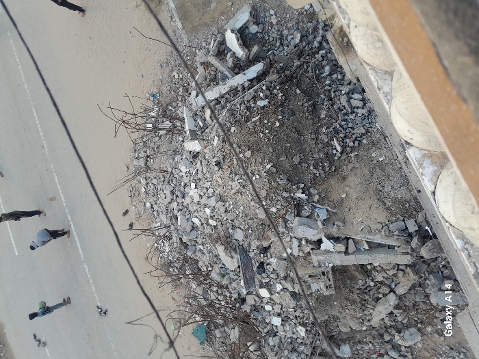 An overhead photograph captures a scene in Khan Yunis, Palestinian Territory, featuring a paved road alongside a substantial pile of building rubble.
In the foreground, a large accumulation of fractured concrete, rebar, soil, and miscellaneous debris occupies the lower half of the image, indicating structural damage or demolition. Twisted metal rods protrude vertically from this rubble. Below the rubble, the edge of a light-colored, possibly concrete, architectural element with rounded top features is visible, suggesting the vantage point is from an elevated structure. Two dark cables traverse the image diagonally from the upper left to the lower right.
The midground transitions from the debris pile to a grey asphalt road marked with two faded white lines. On this road, four individuals are present. To the far left, a person in dark attire holds a green spherical or cylindrical object; a pair of dark shoes lies near them on the asphalt. Adjacent, another individual, dressed in a light grey hoodie and dark pants, is bent forward. A second pair of dark shoes is positioned on the road near this person. Further right, a third person stands upright, wearing dark clothing. The fourth individual is partially visible on the far right, with only their legs and dark pants/shoes apparent. The unpaved area bordering the road appears sandy or dusty.