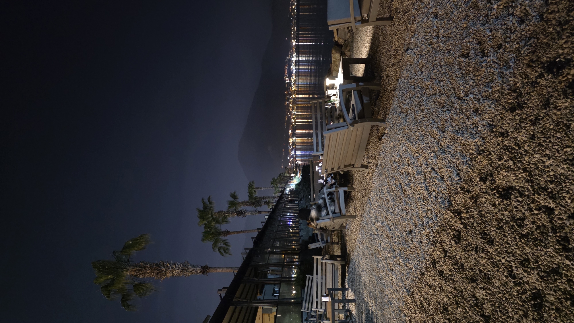 The image captures a serene nighttime scene along a waterfront in Fethiye, Turkey. The view is oriented horizontally, showcasing a tranquil body of water, a distant coastline, and a pebble beach area with seating.
In the mid-ground, a dark mountainous or hilly silhouette rises under a deep blue-black night sky. Below this, a calm expanse of water reflects numerous bright, elongated streaks of light from a distant city or town lining the shore, creating a shimmering effect across the surface.
Running parallel to the water in the middle ground is a paved walkway or promenade, bordered by a line of elegant palm trees. Adjacent to the promenade, a modern building, possibly a restaurant or cafe, features large glass panels that reflect some of the ambient light. Faintly visible on a sign or window pane of this building is the word "hamburg."
To the right, a wide area covered with countless light-colored pebbles extends into the foreground, forming a textured beach or outdoor dining space. Several wooden benches and tables are arranged on this pebbled surface, illuminated by warm artificial lights that cast gentle shadows. One person is clearly visible sitting on a wooden chair at a table, looking downwards, perhaps at a phone or menu. The overall activity suggests a relaxed evening at a waterfront establishment, with guests enjoying the peaceful ambiance and distant city lights.
The time of day is night, with clear weather conditions allowing for crisp reflections and visible details under the artificial lighting. The scene evokes a sense of calm and leisure in a picturesque coastal setting.
