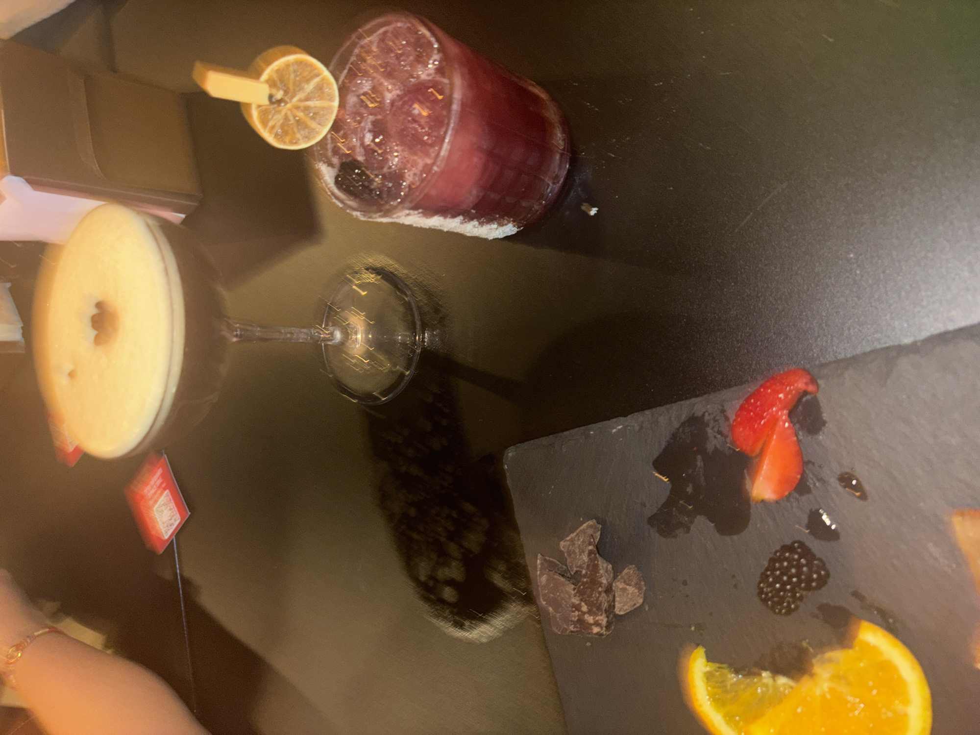 A table in what appears to be a dimly lit bar or restaurant. Three items are arranged: a cocktail in a martini glass with a yellow, round garnish, a red-purple drink in a tall glass with ice, and a slate plate with assorted pieces of fruit and chocolate. A woman’s arm is partially visible in the lower left corner. The restaurant is in Milan, Italy. A small red sign with a QR code sticks out of the martini glass. The scene suggests an evening setting with consumption of alcoholic beverages and snacks.
