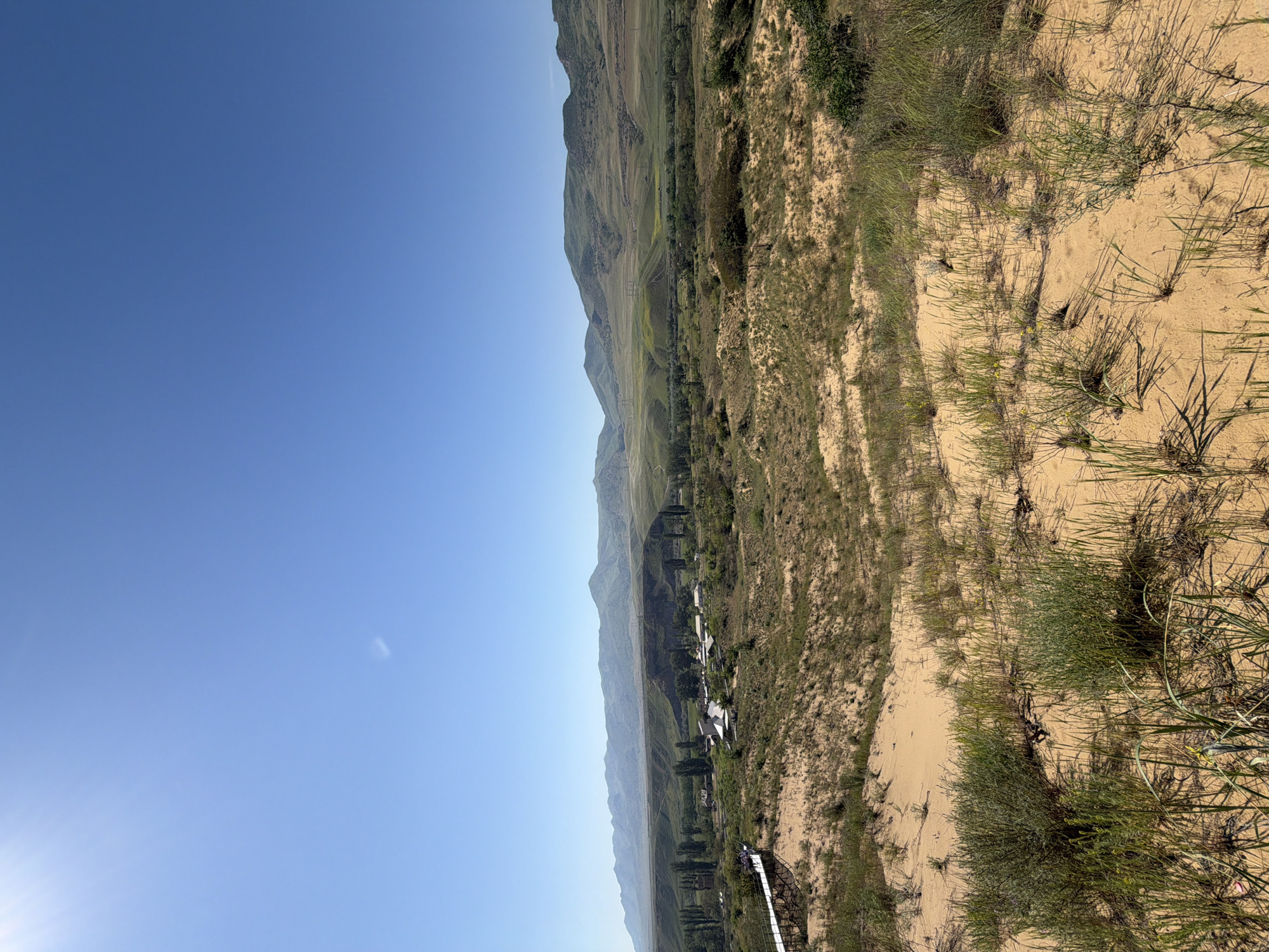 The image depicts a landscape, likely in a mountainous region. The sky is a clear, bright blue. A range of hills/mountains stretches along the horizon. In the foreground, there's sandy terrain with sparse vegetation consisting of grasses and small shrubs. A structure resembling a walkway or bridge is visible in the lower part of the image. A small town is situated between the sandy terrain and the mountains. The precise location within the city of Unknown, Unknown, cannot be determined based on the visual information provided.