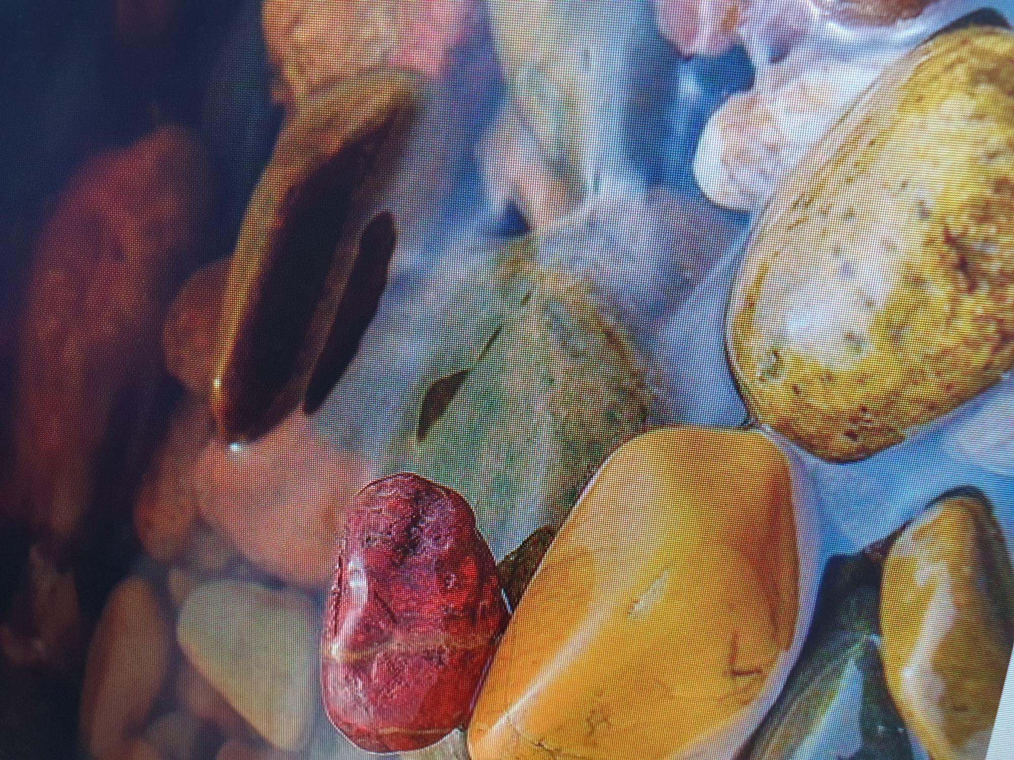The image shows a close-up view of colorful, smooth stones submerged in water, likely in a stream or riverbed. The stones vary in color, with prominent red, yellow, and mottled brown hues. The water is clear, allowing the details of the stones beneath the surface to be visible, and there are some subtle reflections on the water. The overall impression is one of natural beauty and tranquility. This scene could be found in various natural settings within or around Tashkent, Uzbekistan, such as along the Chirchiq River or in parks with decorative water features that utilize natural stones.