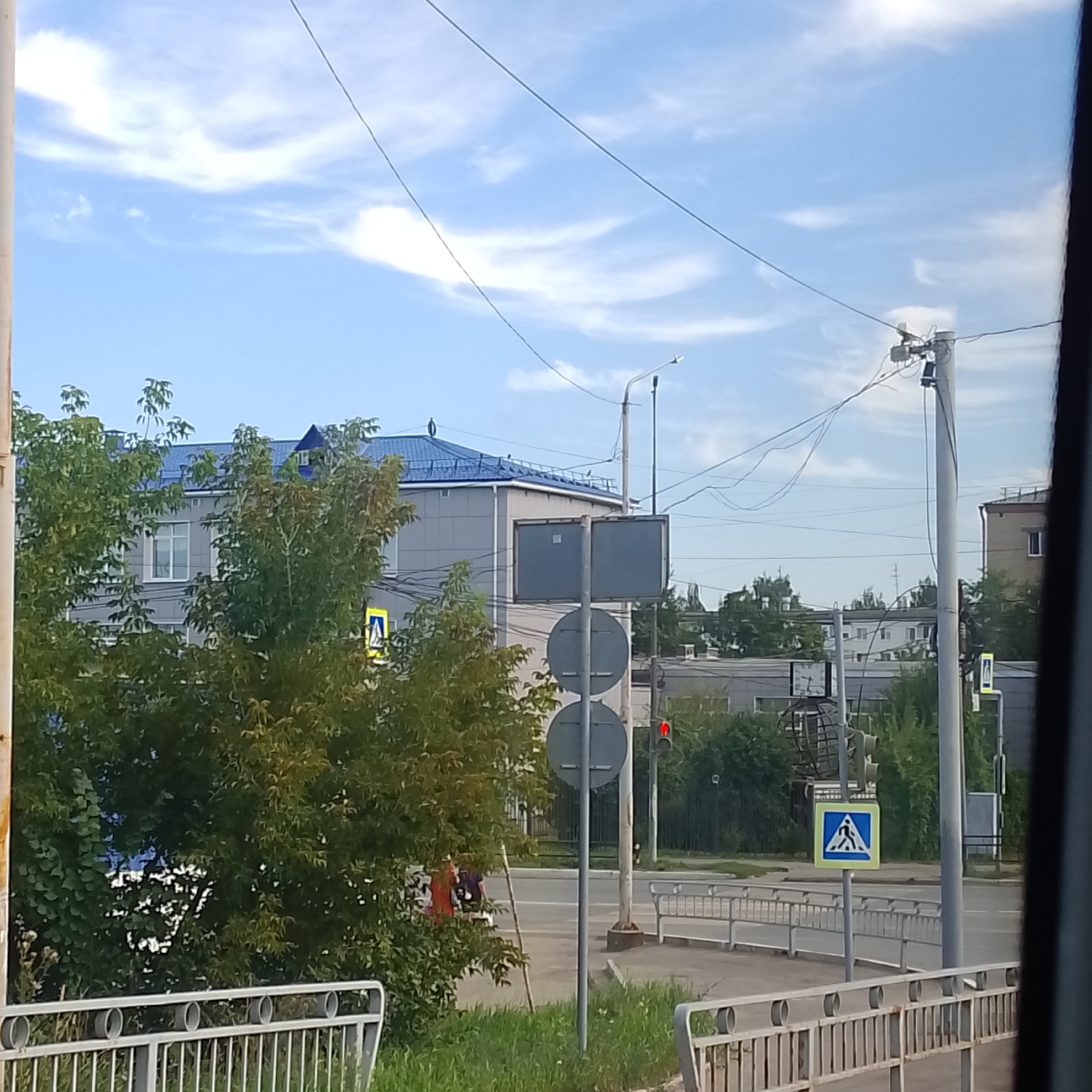 The image shows a view of a street in Chaikovsky, Russia, on a cloudy day. In the foreground, there is a metal fence and green grass. Behind the fence, there is a street with a pedestrian crossing sign and a traffic light showing red. A building with a blue roof and gray walls is visible in the background, partially obscured by trees. Power lines and streetlights are also present. The scene depicts a typical urban environment in Chaikovsky, Russia.