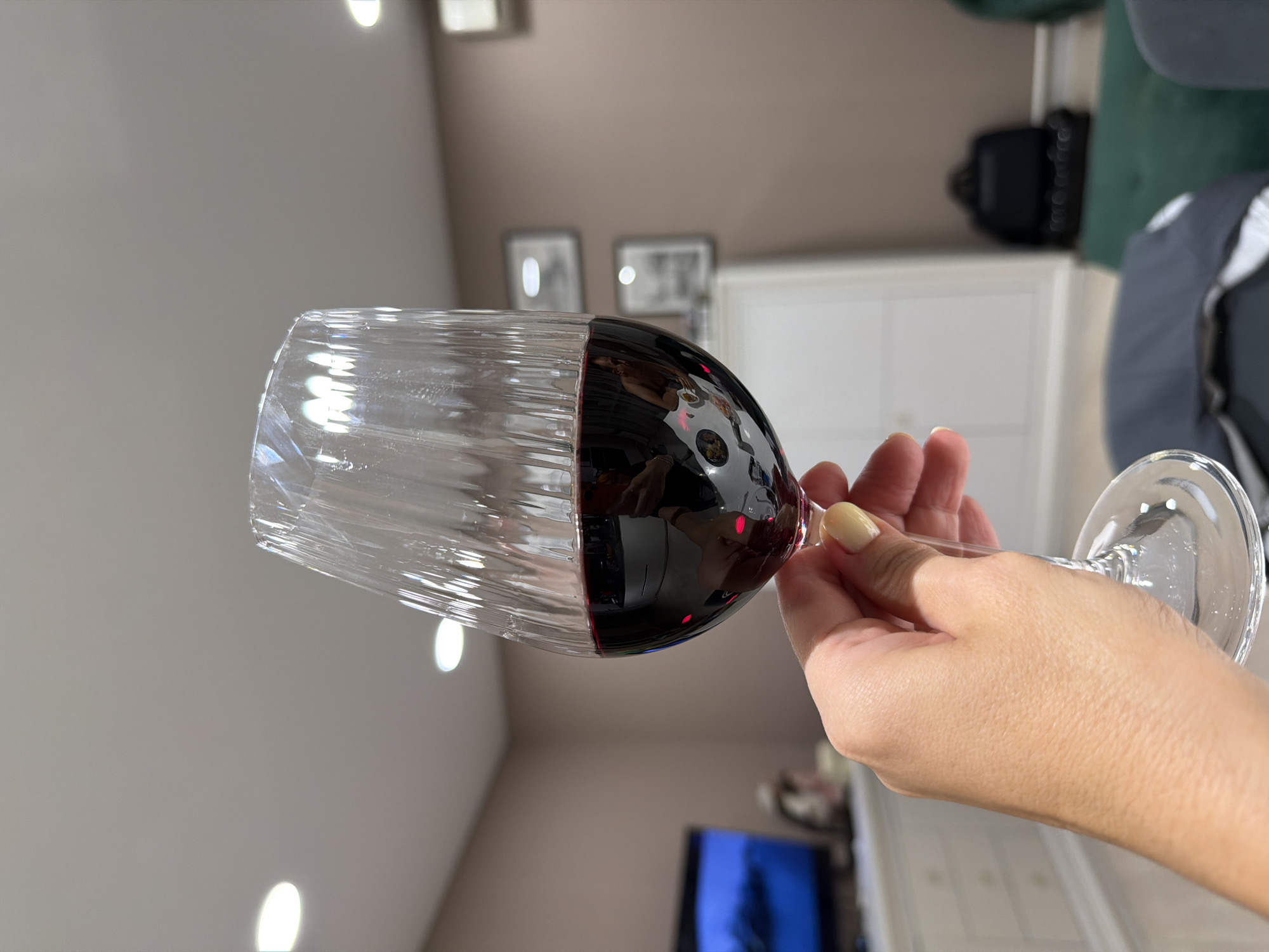 A hand with light-colored nail polish holds a stemmed wine glass filled with red wine. The glass has a textured surface and is positioned in the foreground, slightly off-center to the left. The background appears to be an indoor setting, possibly a living room, with a muted pink wall. A flat-screen television is visible on the left, displaying a blueish image. To the right, there is a white cabinet or dresser, and further back, a black backpack is placed on a green upholstered surface. Two framed pictures are hanging on the wall. The lighting suggests it is indoors, with at least two ceiling lights visible. There is no indication of Buzuluk, Russia in the image.