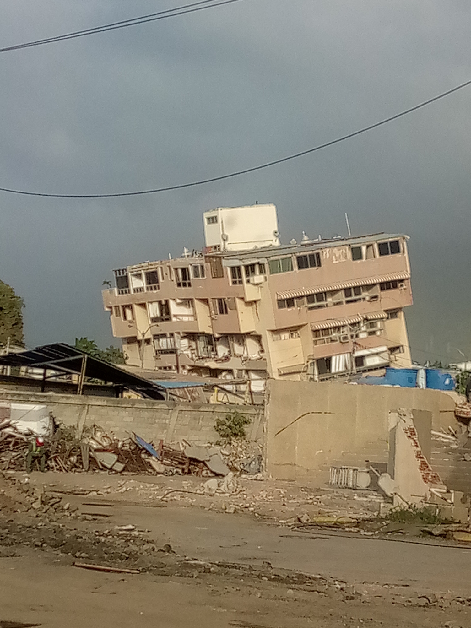 The image depicts a damaged building in what appears to be an urban or suburban environment, with evidence of destruction. The building, a multi-story structure with a light-colored facade, is noticeably tilted, with visible cracks and missing sections. Windows are broken or missing, and awnings are tattered. Debris is scattered around the base of the building and in the foreground, suggesting a recent catastrophic event such as an earthquake, landslide, or bombing. The scene includes a partially constructed or damaged shelter with a dark roof in the middle ground, and a concrete wall that is also showing signs of damage. In the foreground, a pile of rubble includes metal scraps, plastic sheeting, and other construction materials. A single figure wearing a helmet and vest is visible on the left, seemingly surveying the damage or involved in recovery efforts. The sky is overcast and gray. This image likely documents the aftermath of the Vargas tragedy in Caraballeda, Venezuela, which occurred in December 1999, characterized by massive landslides and flooding that caused widespread destruction.