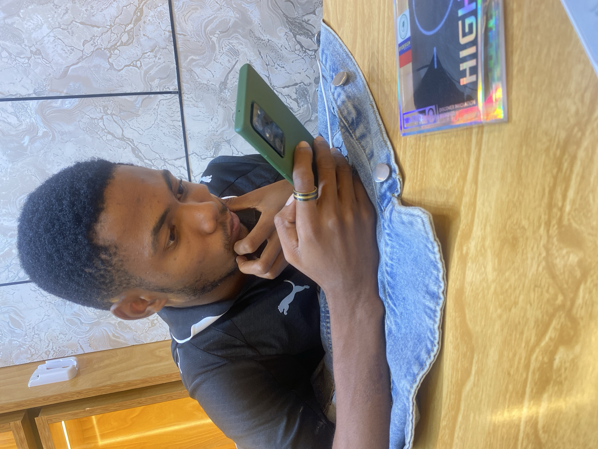 The image shows a man with dark hair and skin looking at a green smartphone. He is wearing a black shirt with a Puma logo and a ring. Next to him is a denim jacket and a package of something with the word "HIGH" on it. The background includes a wood table, a marble-like tiled wall, and what appears to be a white container. I don't have enough information to determine the specific location in Dar es-Salaam.