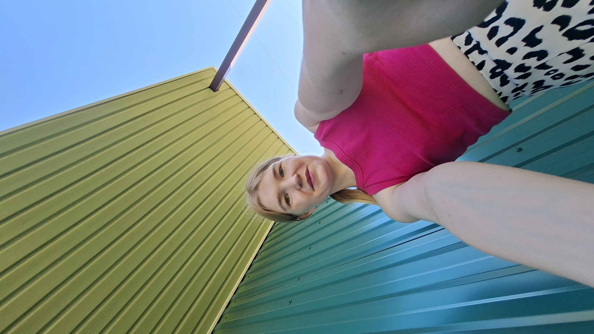 A woman takes a selfie from a low angle, looking upward at the camera. She has blonde hair and is wearing a pink top and animal-print bottoms. Behind her are two corrugated metal walls, one green and one blue. Above the green wall is a blue sky. This photo appears to be taken in Большой Букор, Russia.