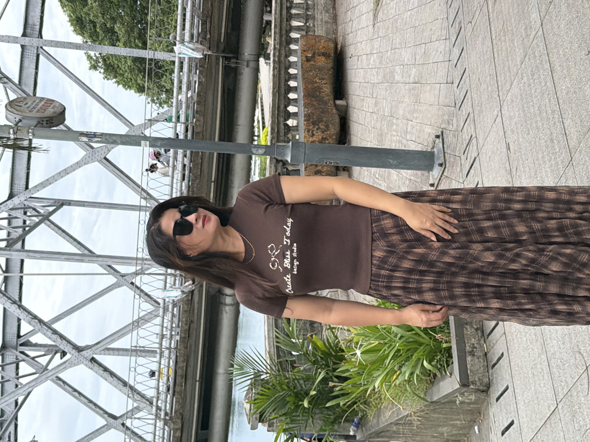 A woman stands on a paved walkway, looking to her right. She wears a brown t-shirt with a white design and text that reads "Create Bliss Today design studio" and a long, pleated brown and tan plaid skirt. She has on black sunglasses. Behind her, a large metal bridge with a complex truss structure spans across a body of water. The walkway has decorative stone railings and planters with green foliage. A lamppost stands to the right of the woman. In the background, people can be seen on the bridge. The scene is set outdoors with a cloudy sky. The location appears to be the bridge in Phu Xuan Ward, Vietnam, a historically significant landmark.