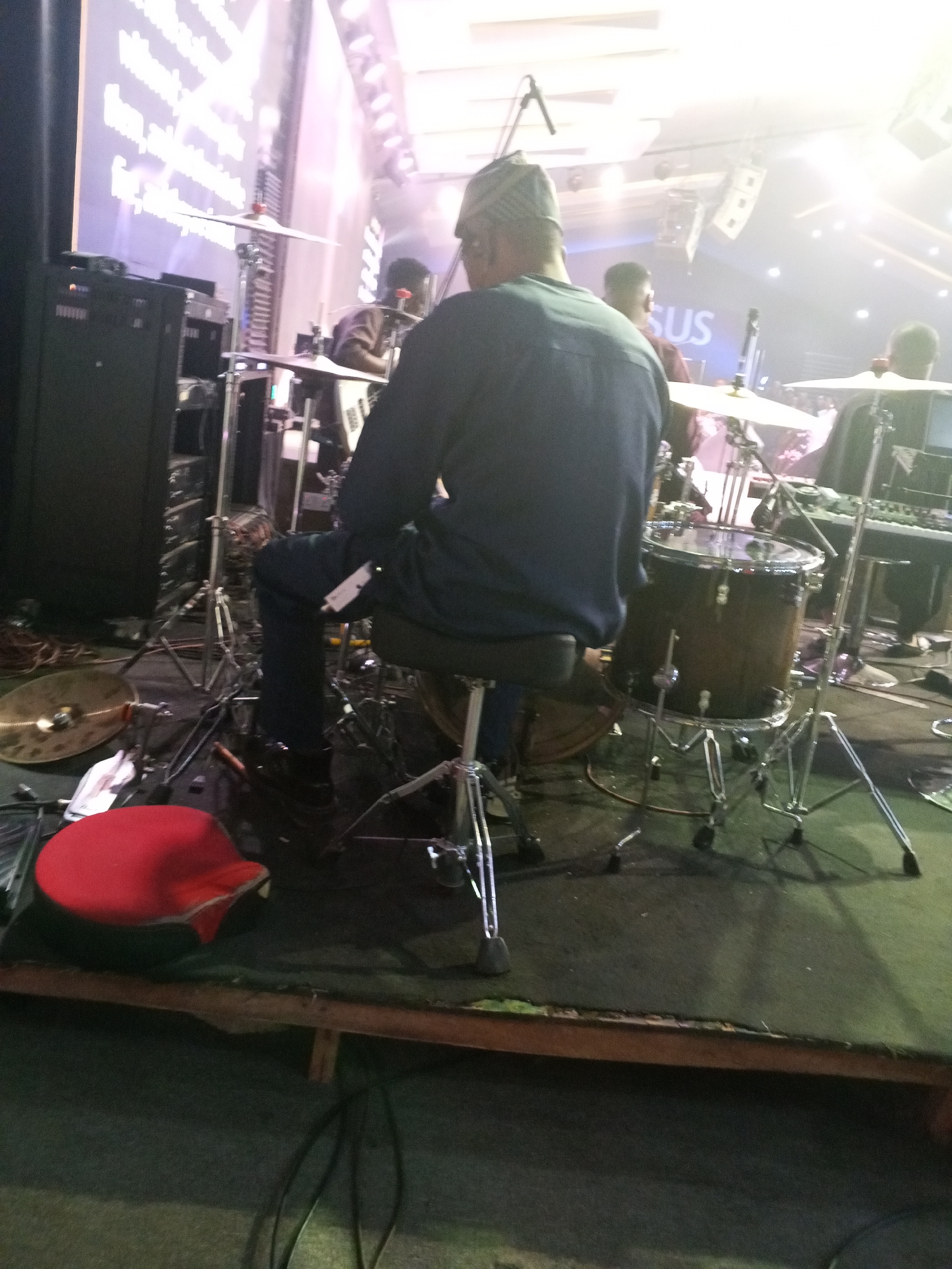 This image captures an indoor stage scene, likely a religious service or gospel concert, at a large venue such as a church auditorium or event center in Abuja, Nigeria. The perspective is from the audience level, looking towards the stage.
In the immediate foreground, a red, somewhat deflated cushion rests on a dark floor surface. Behind it, a raised wooden stage platform, covered in dark green carpet or artificial turf, is visible. The edge of this platform features a brown wooden beam. Electrical cables are present on the floor below the stage.
The central figure is a man seated on a black drum stool, with his back to the viewer. He wears a dark long-sleeved shirt and a patterned cap. His dark trousers and shoes are visible, and he is positioned at a drum kit. To his right, a dark-colored floor tom drum with chrome hardware is clearly visible. Various chrome drum stands and pedals are scattered around the stage floor near his position. A large, brass-colored crash cymbal lies on the stage floor in the bottom left, separate from its stand.
To the left of the main subject, a tall, black audio equipment rack unit with glowing indicators is present. Further left and slightly behind, a large projection screen displays white text, possibly song lyrics or scripture, against a light background.
In the mid-ground, behind the main drummer, another individual is partially visible, also seated and appearing to be a musician, wearing a dark brown or maroon shirt. Further to the right in the mid-ground, a keyboard or synthesizer rests on a stand, with another musician possibly behind it, largely obscured by bright stage lights.
High above the stage, several arrays of white stage lights illuminate the area, creating significant glare and lens flare. A prominent, illuminated sign spelling "JESUS" in capital letters is centrally positioned on the back wall of the stage. Hanging speaker units are also visible high above the stage. The overall environment suggests an active performance or rehearsal setup, with multiple musicians and technical equipment.