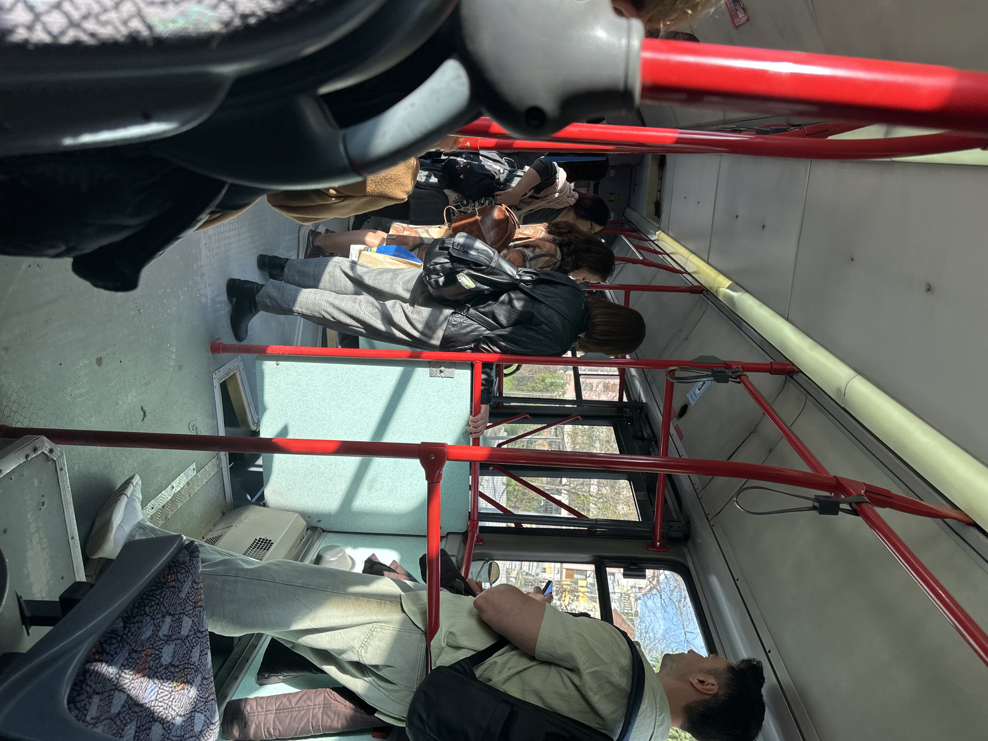 The scene is the interior of a public bus, likely in Sofia, Bulgaria, based on the Cyrillic text on the walls. The bus has red support bars and a gray ceiling. Several passengers are visible. From the front of the bus toward the back, passengers are seated. One passenger is looking at a mobile phone. Another is carrying a dark bag. Further back, other passengers are seated, wearing various jackets and carrying bags. The floor of the bus is light gray. The view through the bus windows shows a city scene with trees and buildings.