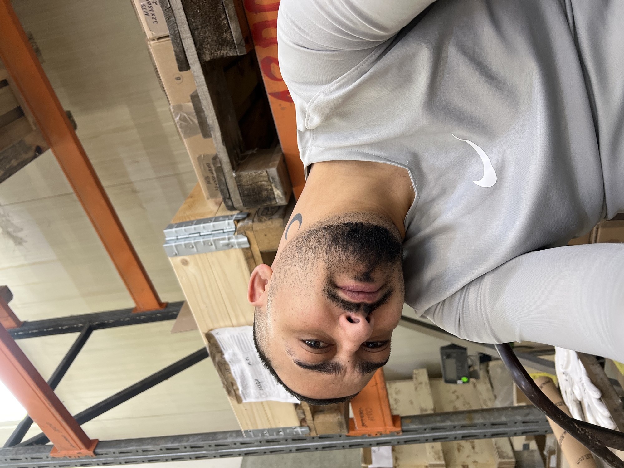 The image is a close-up selfie of a man in an indoor industrial or warehouse environment, captured with a 90-degree clockwise rotation. The man, appearing to be in his 20s or 30s, has short dark hair, a beard, and a goatee. He looks directly at the camera with a neutral expression. A dark, possibly crescent-shaped tattoo is visible on the left side of his neck. He is wearing a light grey, long-sleeved athletic shirt with a white Nike swoosh logo prominently displayed on the chest.
The background reveals typical warehouse elements. Wooden pallets, some stacked with cardboard boxes, are visible above and below the man. A robust metal racking system, featuring orange support beams and grey or black horizontal shelves/supports, extends through the scene. On one of the cardboard boxes, the text "MTS 32X350/0" and "20" is readable, while another box shows "HBS" and partially obscured numbers. In the lower right, white work gloves with the text "WWW.RW..." are faintly visible, alongside a black cable or hose. A small dark electronic device with a green light can be seen on a shelf further back. The lighting suggests an artificially lit interior setting. The location context provided is Sevlievo, Bulgaria.