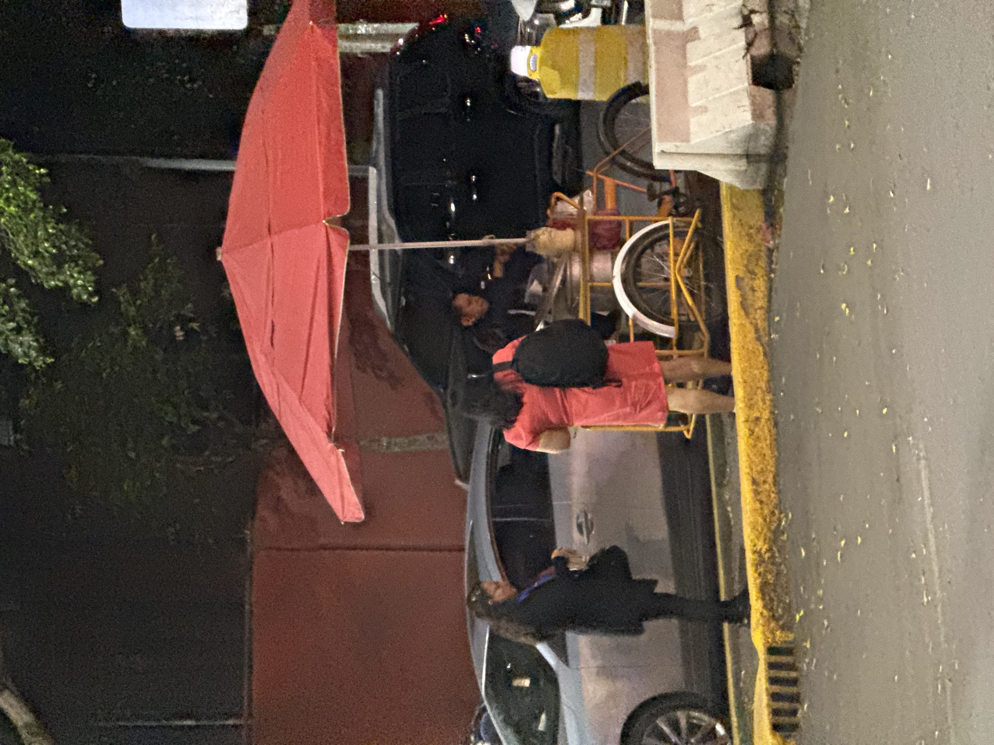 This nighttime image, set in Mexico City, depicts an urban street scene centered around a vendor's stall. A large, partially folded red umbrella dominates the left side of the frame, providing coverage for the stall. Adjacent to it is a yellow-framed vendor's cart, equipped with bicycle wheels and appearing to hold various items, suggesting it's a food or drink stand.
Two individuals are actively engaged near the cart. One person, dressed in a red or orange top and dark pants, stands with their back largely to the viewer, facing the vendor's station. Behind the cart, a second person, likely the vendor, is partially visible in dark clothing, seemingly interacting with the customer. The interaction suggests a purchase or conversation taking place at the stall.
In the background, a dark-colored car is present, and a third individual in dark attire can be seen near it. The scene is illuminated by artificial streetlights, casting a dim glow and creating reflections on the street surface, which also features a yellow curb and a storm drain. A concrete structure or building facade is visible on the right. No discernible text is visible within the image. The overall impression is a bustling yet dimly lit evening street environment.