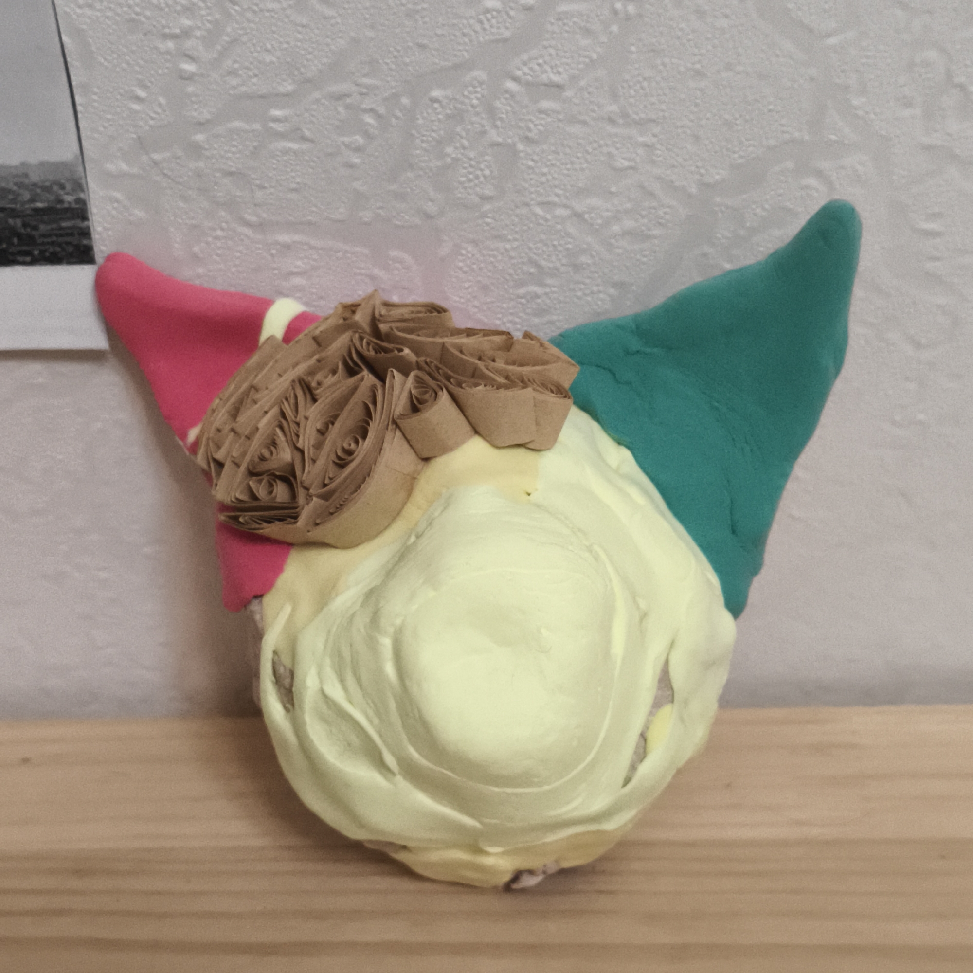 A close-up shot of a colorful, crafted figure with two ears and a cluster of brown paper rolled roses. The ears are pink and green. The main body is covered in a light yellow substance. The figure rests on a wooden shelf. The background consists of a white textured wall and a partially visible photograph.