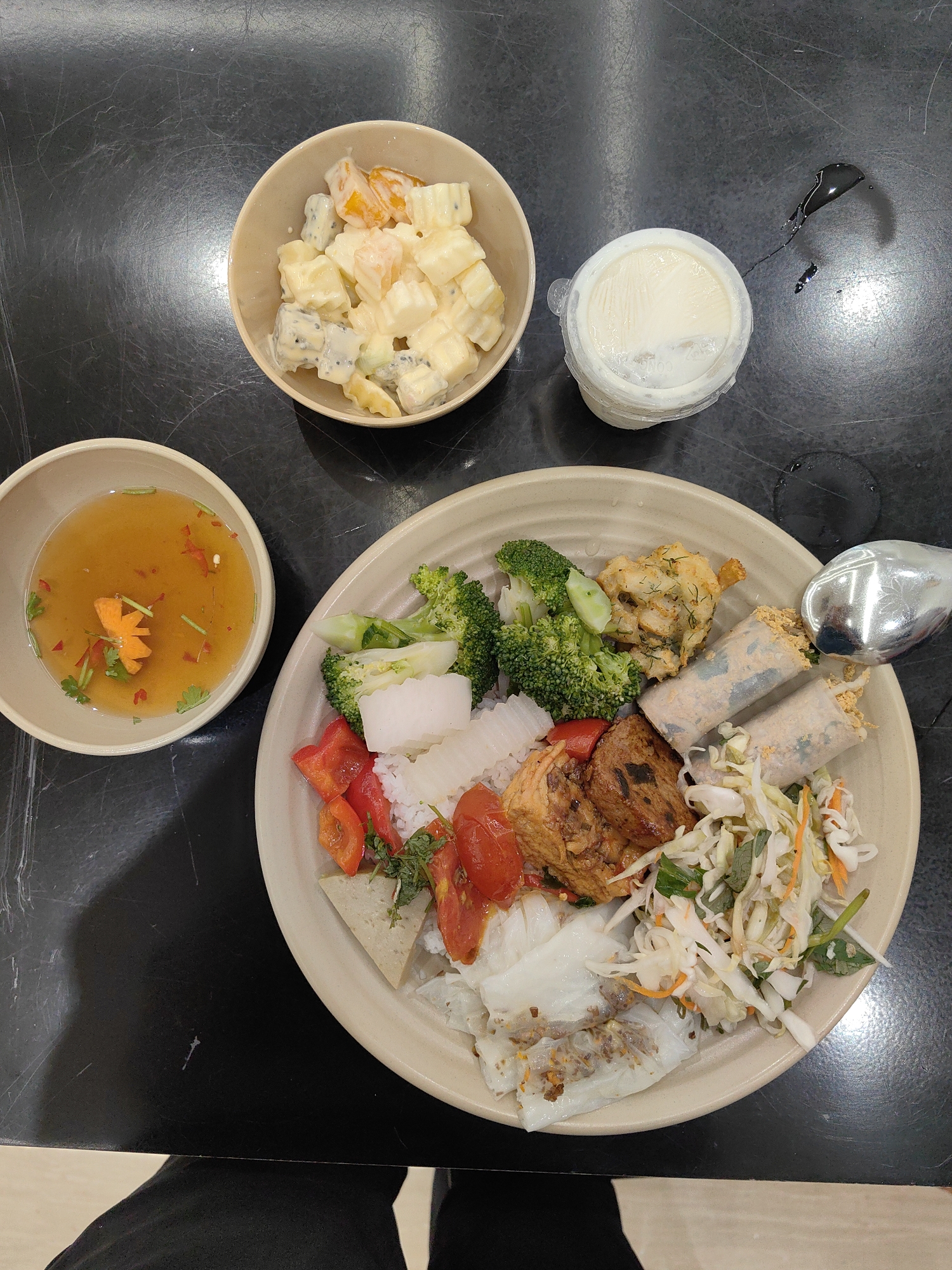 The image shows a meal consisting of Vietnamese food on a black surface. A bowl of fruit salad, a small container of yogurt, and a bowl of dipping sauce are arranged around a larger plate. The large plate contains rice, broccoli, tomatoes, tofu, fried potatoes, salad, and rice paper rolls. A silver spoon rests on the edge of the plate. The setting is likely indoors. The location in the city of Hong Gai Ward, Vietnam is based on the metadata. There are no people visible in the image, although the meal suggests their presence nearby.