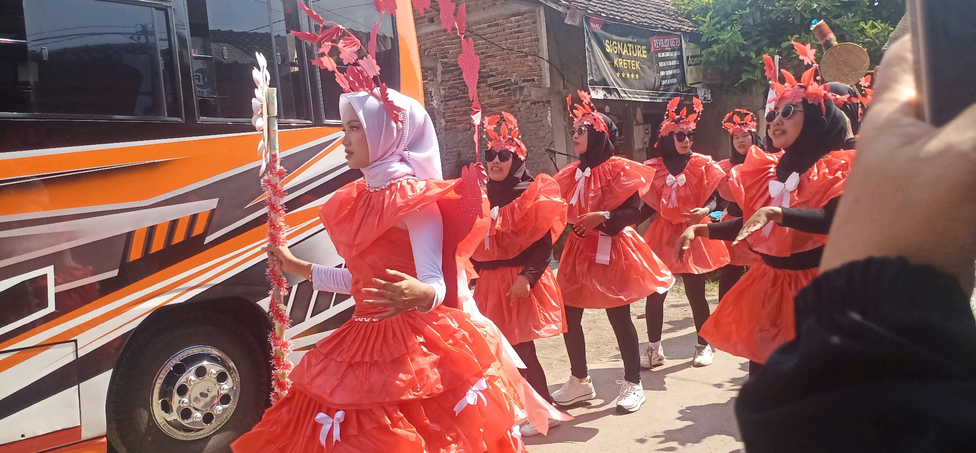 In Kincang, Indonesia, on a sunny day, a group of young women dressed in elaborate orange costumes and red headpieces participate in a parade. The costumes feature voluminous, layered skirts and ruffled neckpieces, adorned with white bows. Most of the women wear headscarves, with some sporting sunglasses. They are positioned in front of a bus with an orange and grey graphic design. The bus has tinted windows and a prominent chrome wheel. Behind the participants, a building with a tiled roof and a brick wall is visible, along with some greenery. A banner on the building reads "SIGNATURE KRETEK". The ground is paved with asphalt. The women appear to be moving in unison, suggesting a coordinated performance or procession. One individual in the foreground is holding a decorated staff with red and white streamers.