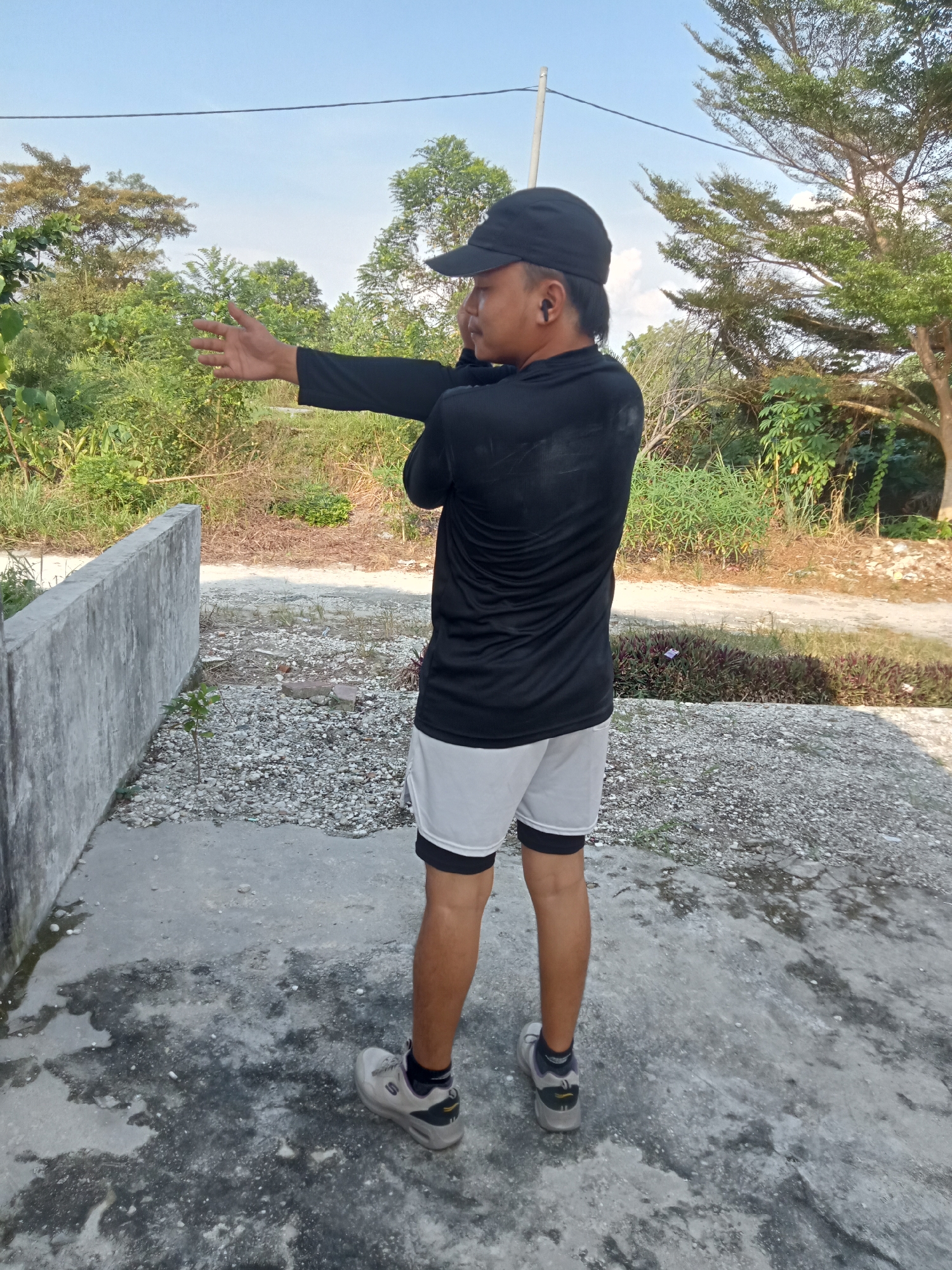 A person wearing a black long-sleeved shirt, white shorts with black compression shorts underneath, a black baseball cap, and gray athletic shoes is stretching their right arm across their chest. They are also wearing black wireless earbuds. The person is standing on a concrete surface with scattered gravel and a low concrete wall to their left. In the background, there are various trees and vegetation, suggesting an outdoor environment. The scene appears to be in Kualu, Indonesia, likely a residential or semi-rural area, given the presence of a concrete wall and the surrounding natural elements. The sky is clear and blue, indicating daylight.