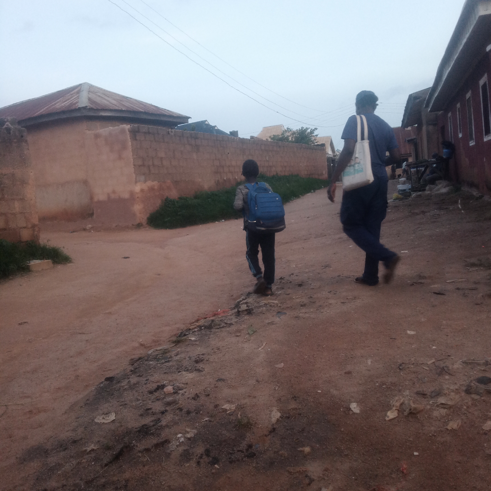 In Jos, Nigeria, a young person wearing a blue backpack walks away from the camera on a dirt road. Behind them, an adult also walks away, carrying a white tote bag over their shoulder. They are in a semi-urban area with buildings made of brick and mud. The sky is overcast.