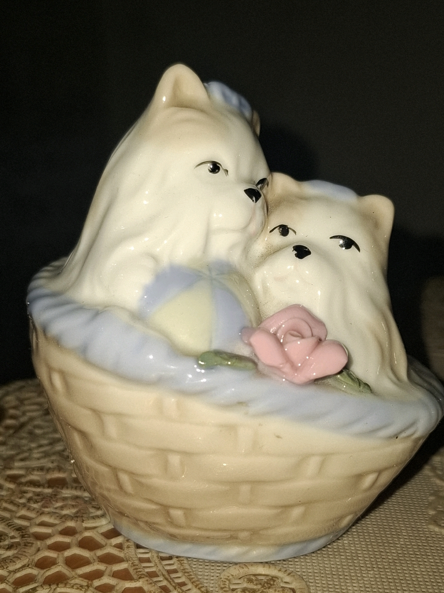 This is a close-up, eye-level photograph of a ceramic figurine of two white puppies nestled in a light brown basket. The puppies have black eyes and noses, and the larger puppy on the left is leaning its head against the smaller puppy on the right. A pink rose with green leaves rests in front of the puppies. The basket has a textured, woven appearance and a light blue rim. The background is dark and out of focus. The figurine is placed on a textured, beige surface. The image does not contain any information about the city of Almaty, Kazakhstan.