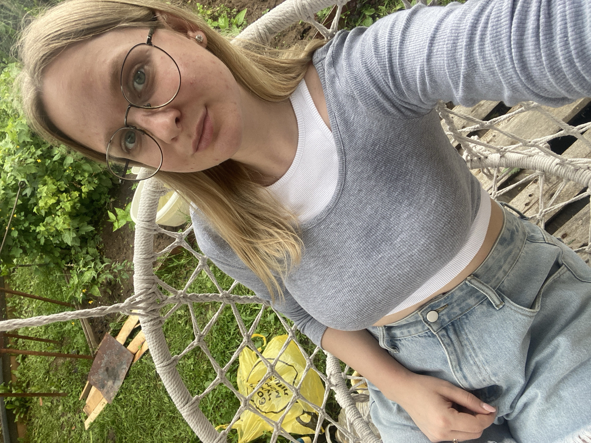A young woman with blonde hair and glasses is sitting on a rope-strung chair outdoors in what appears to be a rural or garden setting. She is wearing a grey long-sleeved crop top over a white undershirt and light-wash denim jeans. The background consists of greenery, including trees and grass, with some wooden planks and a yellow plastic bag visible on the ground. The scene suggests a casual, relaxed atmosphere, possibly a backyard or a park. The location of Kauchuk Stantsiya, Russia, is not explicitly evident from the visual information alone.