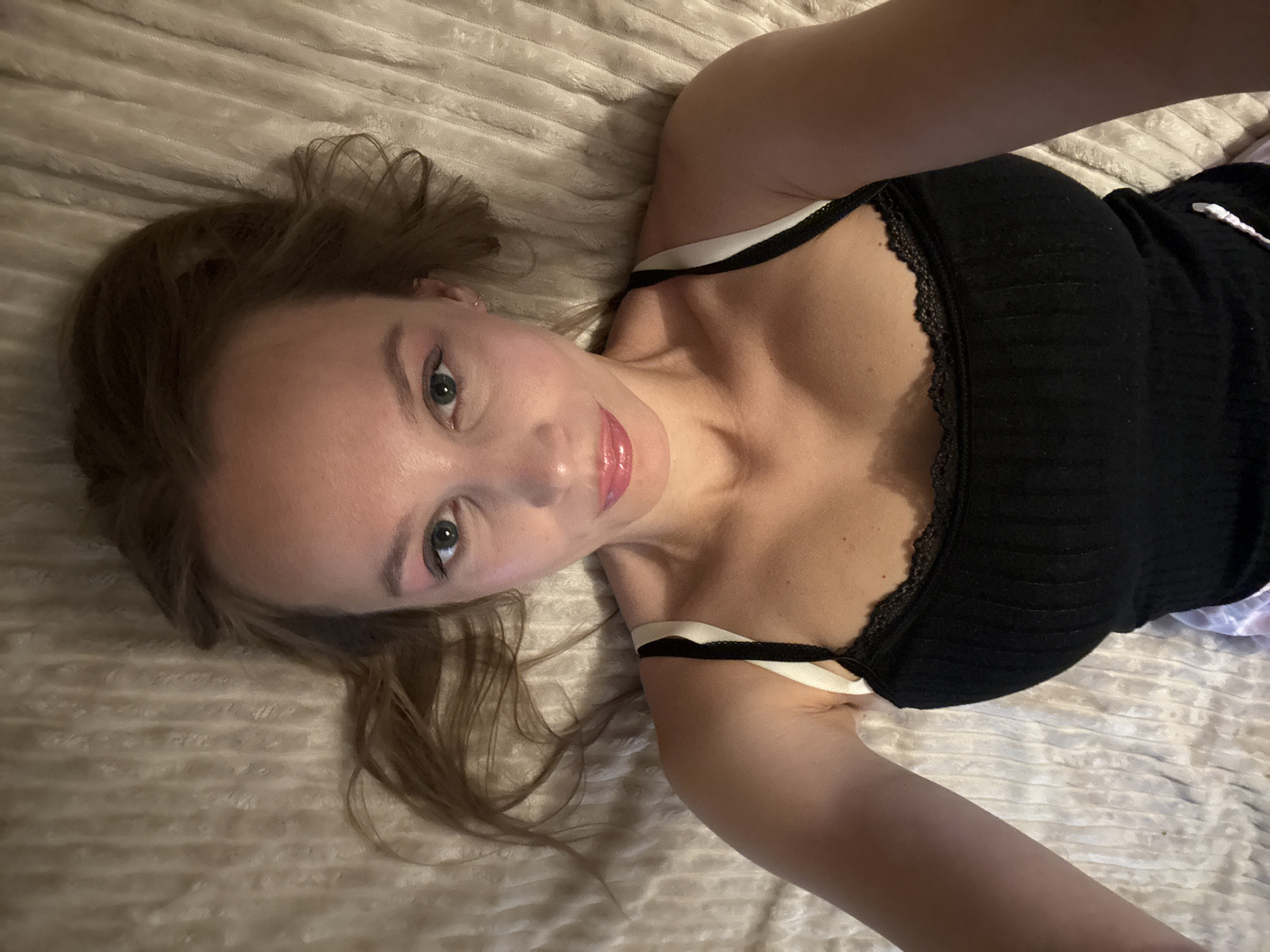 A woman lies on a textured, light-colored bedspread, looking upwards at the camera. Her hair is a light brown and is spread out around her head. She is wearing a black tank top with white straps and a lace trim. Her makeup includes pink eyeshadow and lip gloss. Her face is tilted slightly to her left. The image is taken from a high-angle perspective. The scene appears to be indoors.