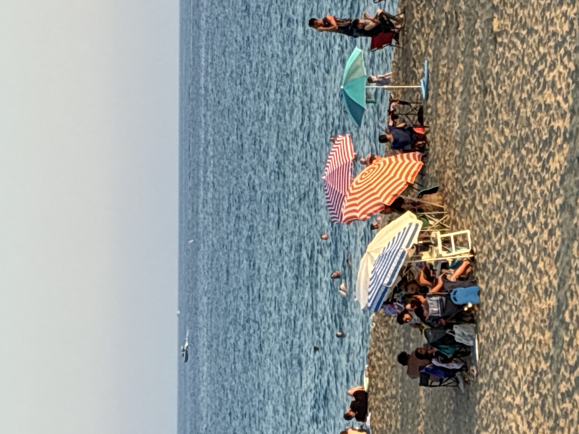 The image depicts a crowded beach scene in Alanya, Turkey. Several people are relaxing on the sand, seated in chairs and under umbrellas. The water is a vibrant blue, with gentle waves suggesting calm weather. Multiple beach umbrellas, including a striped red and white one and a blue and white striped one, provide shade. People are engaged in various beach activities such as sunbathing and socializing. The time of day appears to be late afternoon, as indicated by the warm, golden light casting shadows on the sand. The overall atmosphere is one of leisure and enjoyment. No visible text is present in the image.