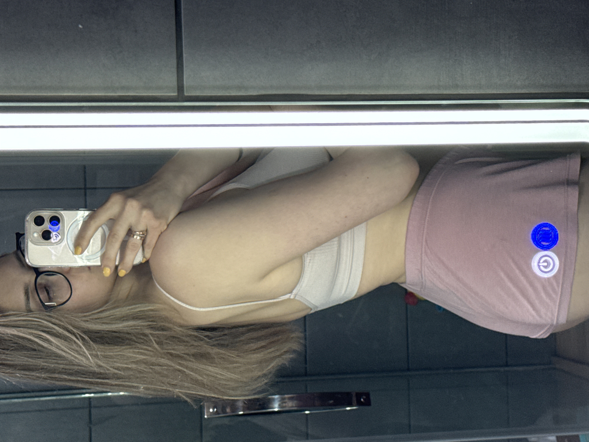 A woman is taking a selfie in a bathroom mirror. She has blonde hair, glasses, and is wearing a light-colored sports bra and pink shorts. Her nails are painted yellow. The background shows grey tiled walls and a bright, horizontal light fixture. There are blue and white circle details on the bottom of her shorts. It is unclear where in Chaikovsky, Russia this image was taken.