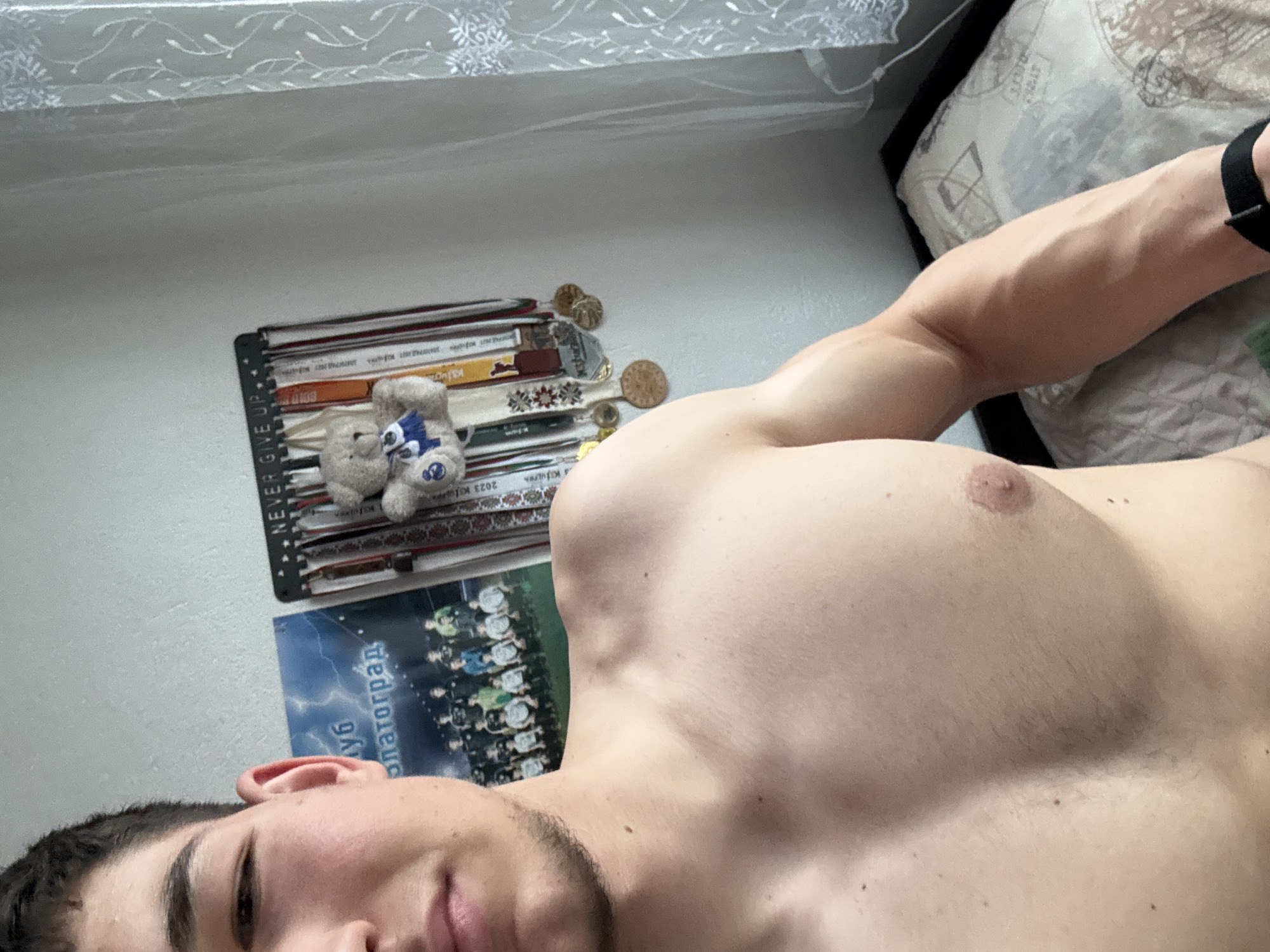 A close-up selfie captures a shirtless young man, visible from the mid-chest up, lying down and looking towards the camera with a neutral expression. His muscular right arm, chest, and a portion of his face with stubble are prominent. A black wristband is worn on his right wrist.
In the background, on a light-colored wall, a black display rack is adorned with numerous medals and colorful ribbons. Two small, light-colored stuffed bears, one with blue accents, sit on the rack among the medals. Below the medal rack, a poster is partially visible, showing a group of people, likely a sports team, in uniform. Text on the poster clearly reads "Златоград" (Zlatograd) and "клуб" (club) in Cyrillic, along with "2023". Further text "NEVER GIVE UP" is partially visible on the medal display rack itself, as is "2023".
Behind the man's arm and head, a bed with a patterned pillow or duvet is visible. In the upper left corner of the frame, a white, sheer curtain with a delicate pattern can be seen, suggesting the presence of a window. The lighting indicates it is daytime. The scene depicts an indoor setting, likely a bedroom in Zlatograd, Bulgaria, showcasing personal achievements and belongings.