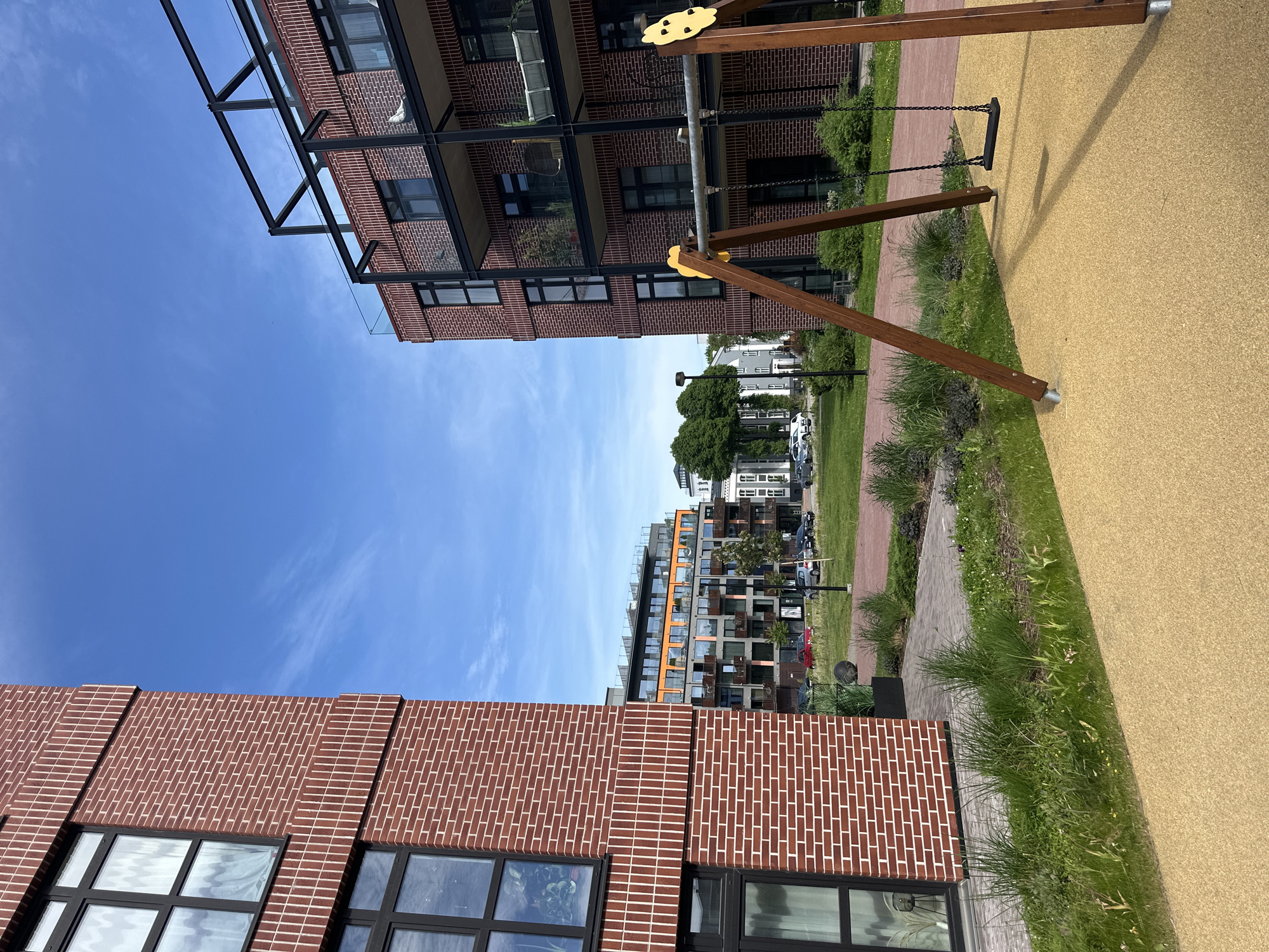 The image depicts a modern residential complex in Tallinn, Estonia. In the foreground, a playground swing set with wooden supports and black seats is visible on a yellow rubberized surface. To the left, a red brick building with large black-framed windows dominates the scene. Further into the background, another apartment building with a more varied facade, including orange accents and balconies, stands under a clear blue sky with scattered white clouds. Several cars are parked on a paved area between the buildings and a strip of green lawn. The overall impression is one of a contemporary urban environment with amenities for residents.