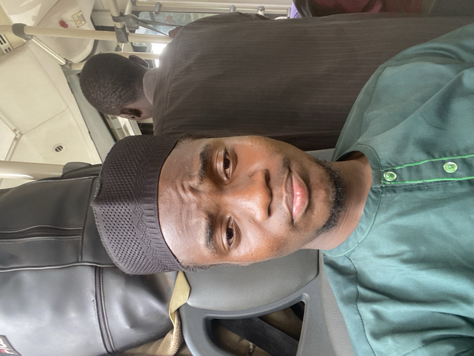 Inside a vehicle, likely a public transit bus in Epe, Nigeria, a young man of dark complexion is depicted in a medium close-up, facing the camera directly. He wears a dark green traditional long-sleeved top with a band collar and light green buttons/trim, along with a black, textured cap covering his head. His expression is neutral with slightly furrowed brows. Behind him, a grey Fila-branded backpack rests on a seat. To his right and slightly behind, another individual with short dark hair is partially visible from the rear, wearing a dark brown garment and holding onto a white overhead grab bar. The vehicle's interior features a white ceiling with ventilation grilles, white vertical and horizontal grab bars, and grey upholstered seats.