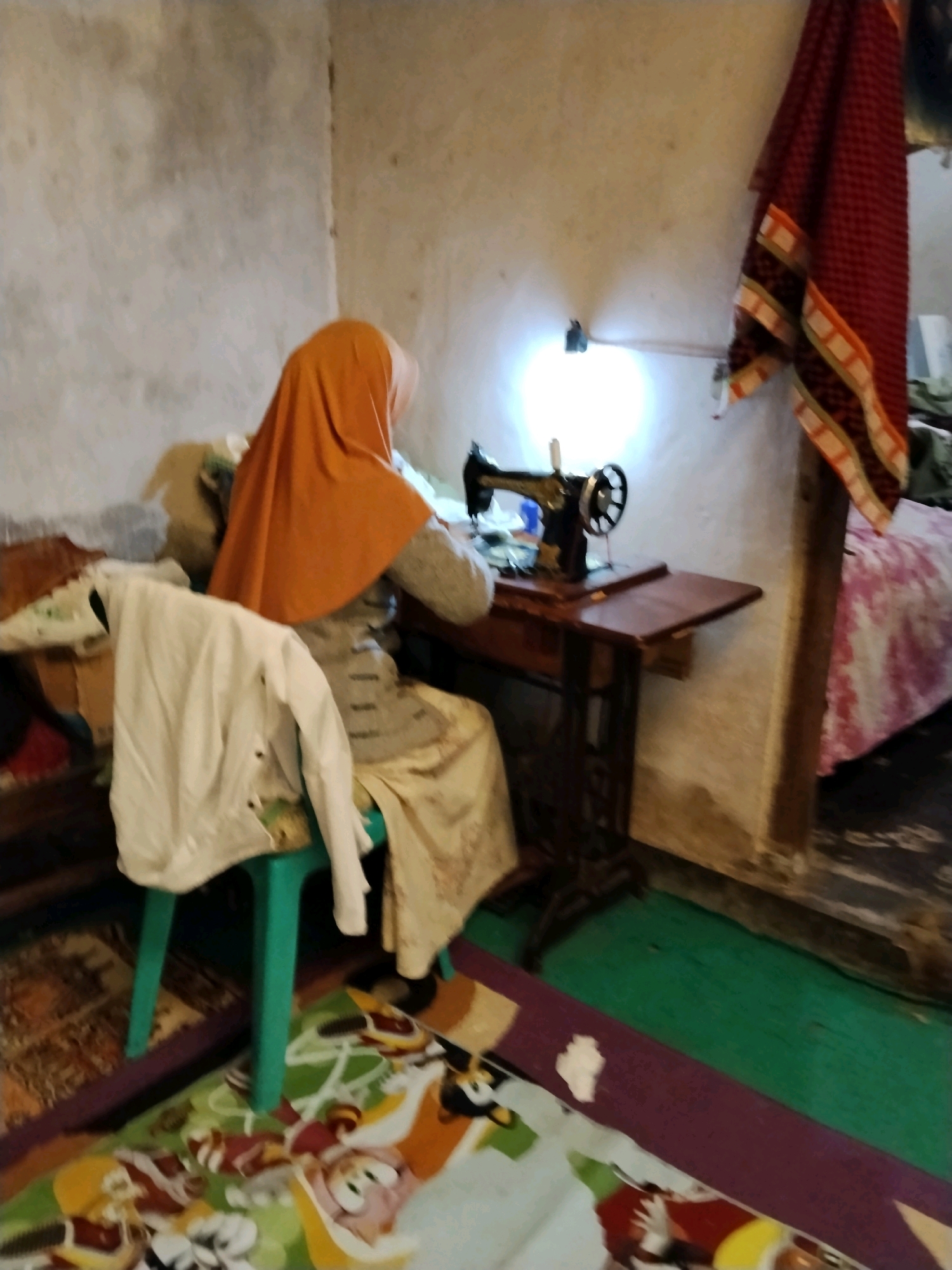 In a dimly lit room with a concrete wall and floor, a person wearing an orange hijab and a patterned garment sits at a vintage sewing machine. The sewing machine is on a dark wooden table with drawers. To the left of the sewing machine, a white shirt is draped over a teal stool. A red and black checkered textile with an orange and white striped border hangs on the wall to the right of the sewing machine. A bed with a pink patterned cover is visible in the background on the right. The room appears to be located in Panggungan, Indonesia.