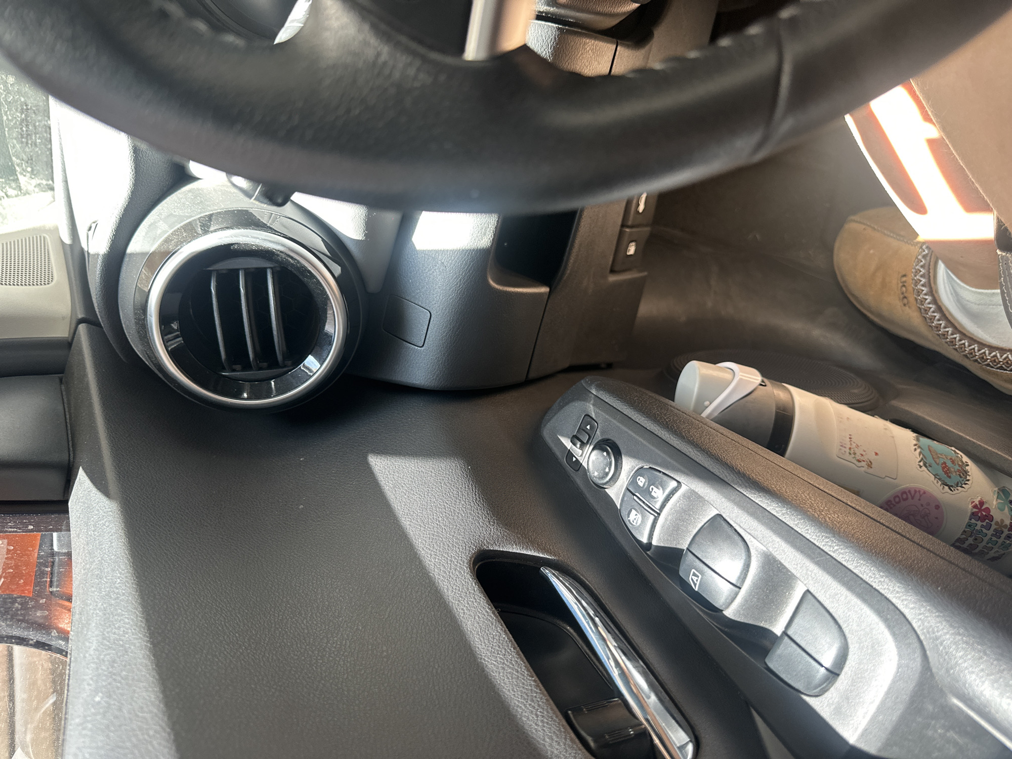 The image provides an interior view of a car, observed from the driver's perspective, looking towards the passenger side and down. The scene is illuminated by bright, direct sunlight streaming into the vehicle, creating strong highlights and distinct shadows, suggesting a sunny daytime in Jesús del Monte, Mexico.
A section of a black, textured steering wheel occupies the top of the frame. Below it, on the dashboard to the left, is a circular air vent with a metallic trim and vertical slats. The dashboard and door panels are made of black, textured plastic.
Prominently featured on the door panel is a control module for various functions. It includes buttons for power windows, one marked with an "A," suggesting an automatic function, and others for door locks, indicated by lock and unlock symbols. A joystick-like control for mirror adjustments is also visible. Below these controls, a gleaming silver door handle is partially seen.
On the passenger side floor, a person's foot is visible, clad in a light brown, moccasin-style slipper. The "UGG" logo can be faintly discerned on the side of the shoe. Adjacent to the shoe, a white bottle or tumbler, decorated with several colorful stickers, lies on the floor. Visible text on these stickers includes "GROOVY" and part of another phrase appearing as "JOY WITH," accompanied by various floral and abstract designs.
No particular activity or event is depicted, other than the car being occupied. The overall impression is a casual, everyday moment inside a vehicle.