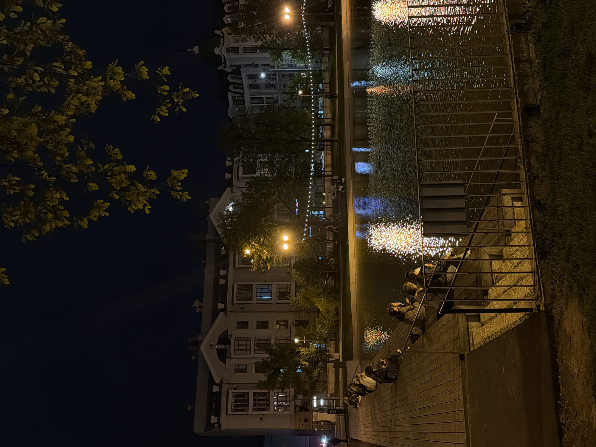 The image depicts a nighttime scene in Kazan, Russia, likely near the Qaban Lakes area. Illuminated buildings are visible along the waterfront, with reflections shimmering on the water's surface. Strings of lights stretch along the water’s edge, reflecting as points of light. People are sitting on a waterfront embankment, some leaning against railings, observing the lake and city view. A tree with illuminated leaves frames the left side of the image, while a patch of grass is visible on the right edge. The scene is dark, with artificial lights creating contrast and defining the urban environment.