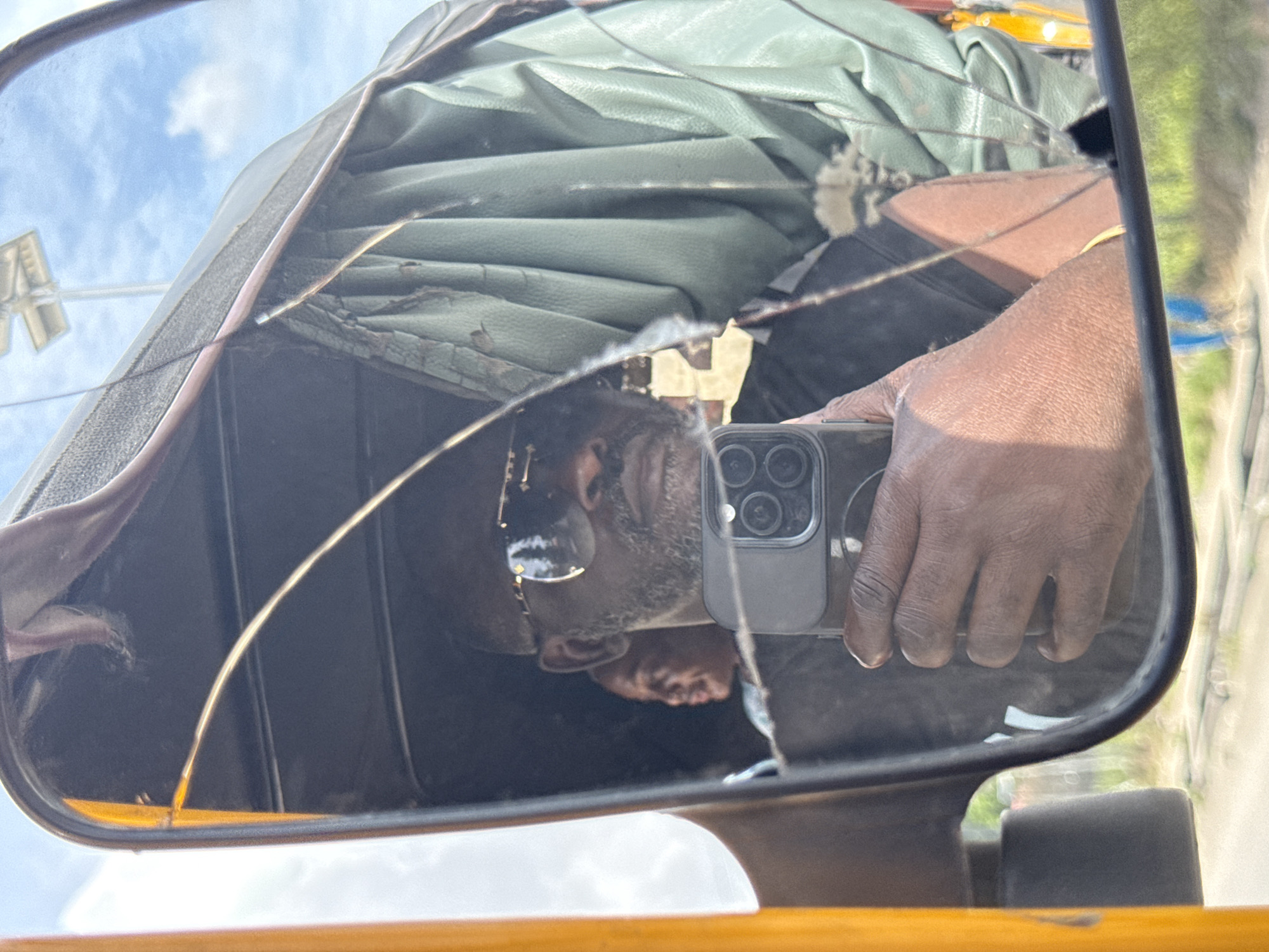 A fractured rearview mirror of a yellow tricycle (Keke Napep) reflects a man holding a smartphone. The mirror's damage obscures portions of the man's face and the interior of the vehicle, which has a dark green, possibly leather, roof. The smartphone held by the man has multiple camera lenses visible. Outside the vehicle, light-colored structures and sky are visible. Based on the description, it is implied to be in Ado, Nigeria.