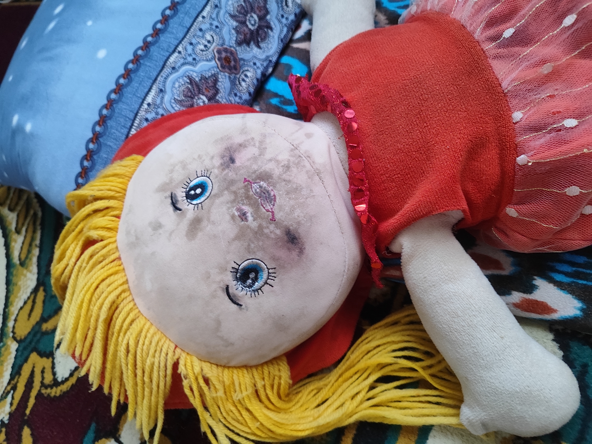 A stuffed doll lies on its back on a patterned surface, possibly a blanket or rug. The doll has blonde yarn hair and a light-colored fabric face with one visible blue eye and a faint smile. A red fabric garment covers the doll's torso, and a sheer, red, polka-dotted skirt is attached to the garment. The doll's left arm is visible and appears to be made of white fabric. The background includes a blue pillow with white polka dots and a decorative blue and patterned fabric. The scene is indoors and appears to be in a residential setting.