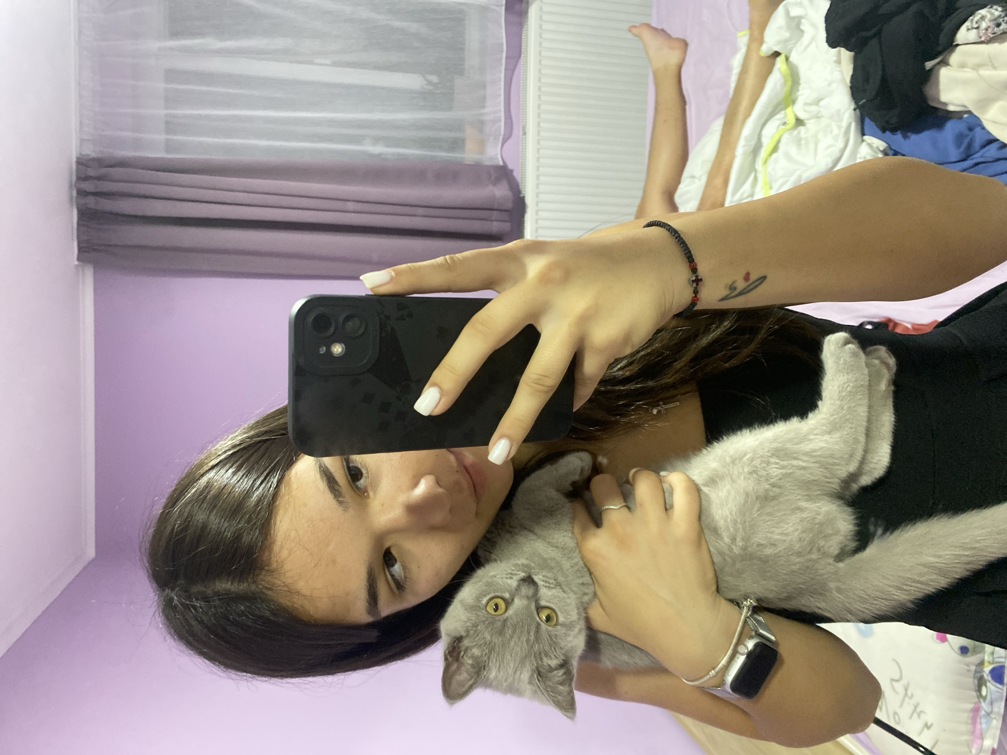 A young woman with long, dark hair takes a selfie with her phone while holding a light gray kitten. She is wearing a black tank top and a silver watch. The kitten is looking at the camera with wide yellow eyes. In the background, a bed with white and yellow linens is visible, with a person's legs lying on it. The room has lavender walls and purple curtains covering a window. A white radiator is below the window.