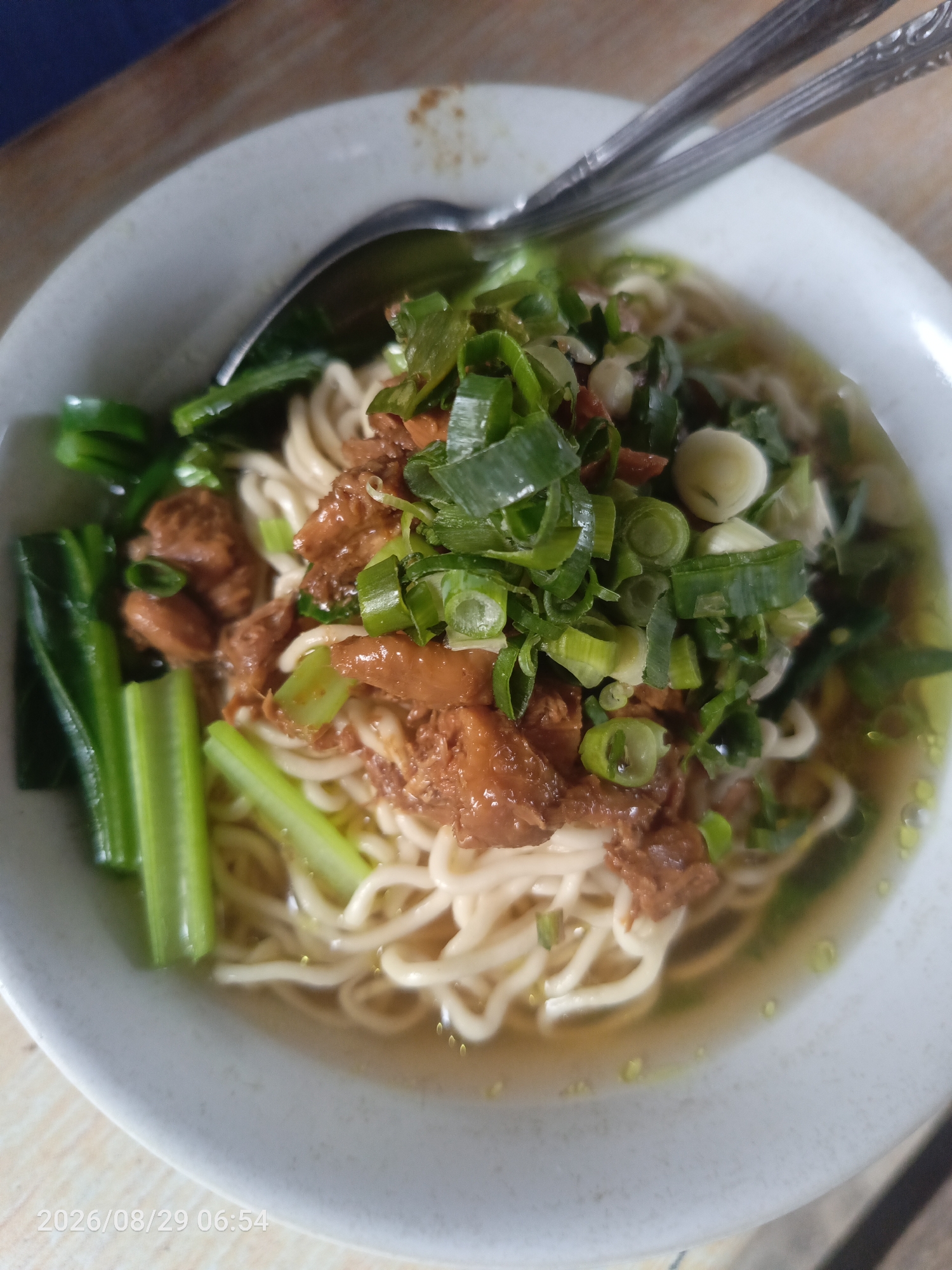 A close-up, overhead shot features a bowl of chicken noodle soup. The soup is in a white bowl with a subtle texture. The noodles are thin and white, submerged in a light-colored broth. Topped with shredded chicken, chopped green onions, and what appear to be small pieces of garlic. On the left side of the bowl are several stalks of green leafy vegetables, possibly bok choy. Two silver utensils, a spoon and a fork, are placed across the top of the bowl. The background consists of a wooden surface. A timestamp in the bottom left corner reads "2026/08/29 06:54". The image is taken in Sangiang, Indonesia.