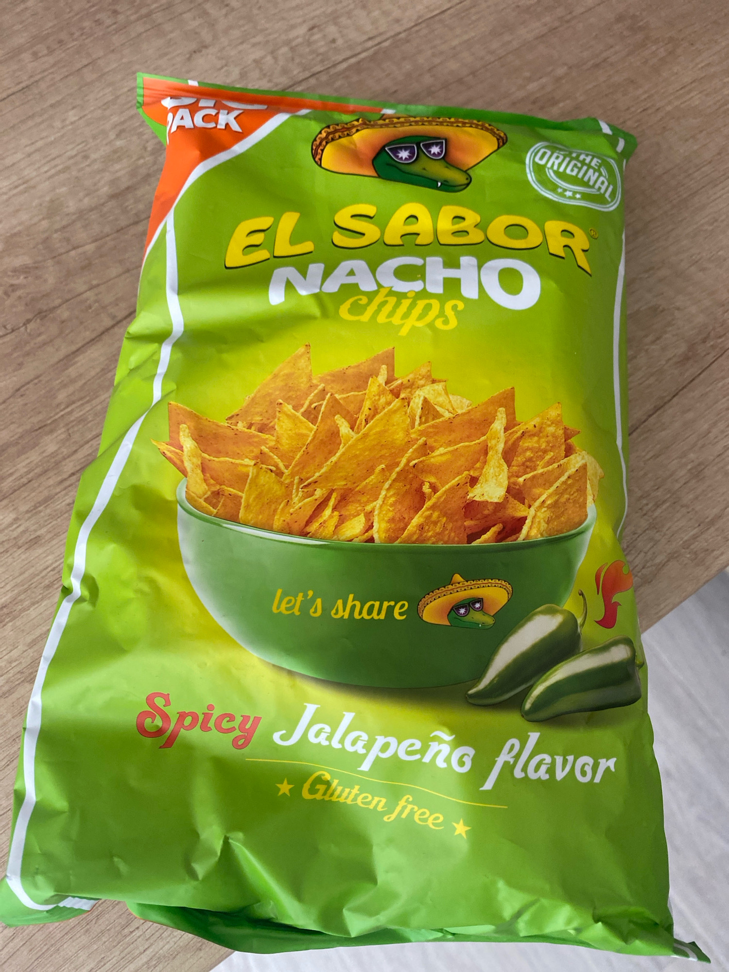 A bright green bag of "El Sabor Nacho Chips" in "Spicy Jalapeño flavor" is displayed on a light brown wooden surface. The bag is partially open at the top, revealing a pile of golden-yellow tortilla chips. A cartoon alligator wearing a sombrero and sunglasses is depicted on the upper portion of the bag, with a circular emblem reading "THE ORIGINAL" to its right. Below the chips, a green bowl with the text "let's share" is visible, accompanied by two jalapeño peppers. The bottom of the bag prominently features the text "Spicy Jalapeño flavor" in a stylized font, with "Gluten free" written below in yellow. The bag has some minor creases and wrinkles, indicating it is not perfectly flat. The lighting suggests it is an indoor setting, likely a home or store. There is no information available to confirm its location in Chișinău, Moldova.