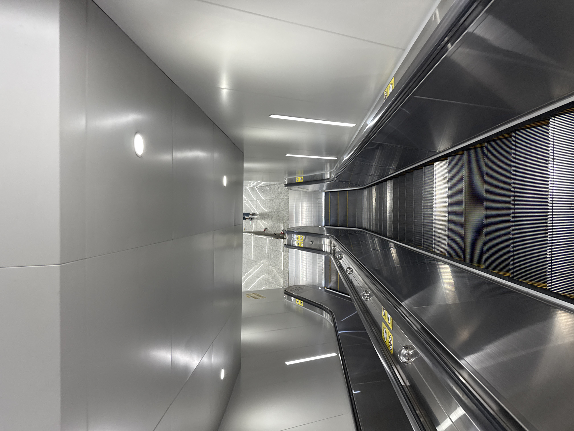 An outdoor escalator in Haizhu District, China, descends from a brightly lit, modern structure with a white, angled ceiling. The escalator itself is made of stainless steel, with a continuous handrail and a series of metal steps. Yellow safety markings are visible on the edges of the steps. Several vertical, linear lights are embedded in the white wall to the right of the escalator. At the bottom of the escalator, the ground appears to be a patterned tile floor. Two individuals, dressed in casual clothing, are visible at the top of the escalator, one walking away from the camera and the other further in the distance. The overall scene is clean and contemporary, indicative of a public transportation hub or modern building.