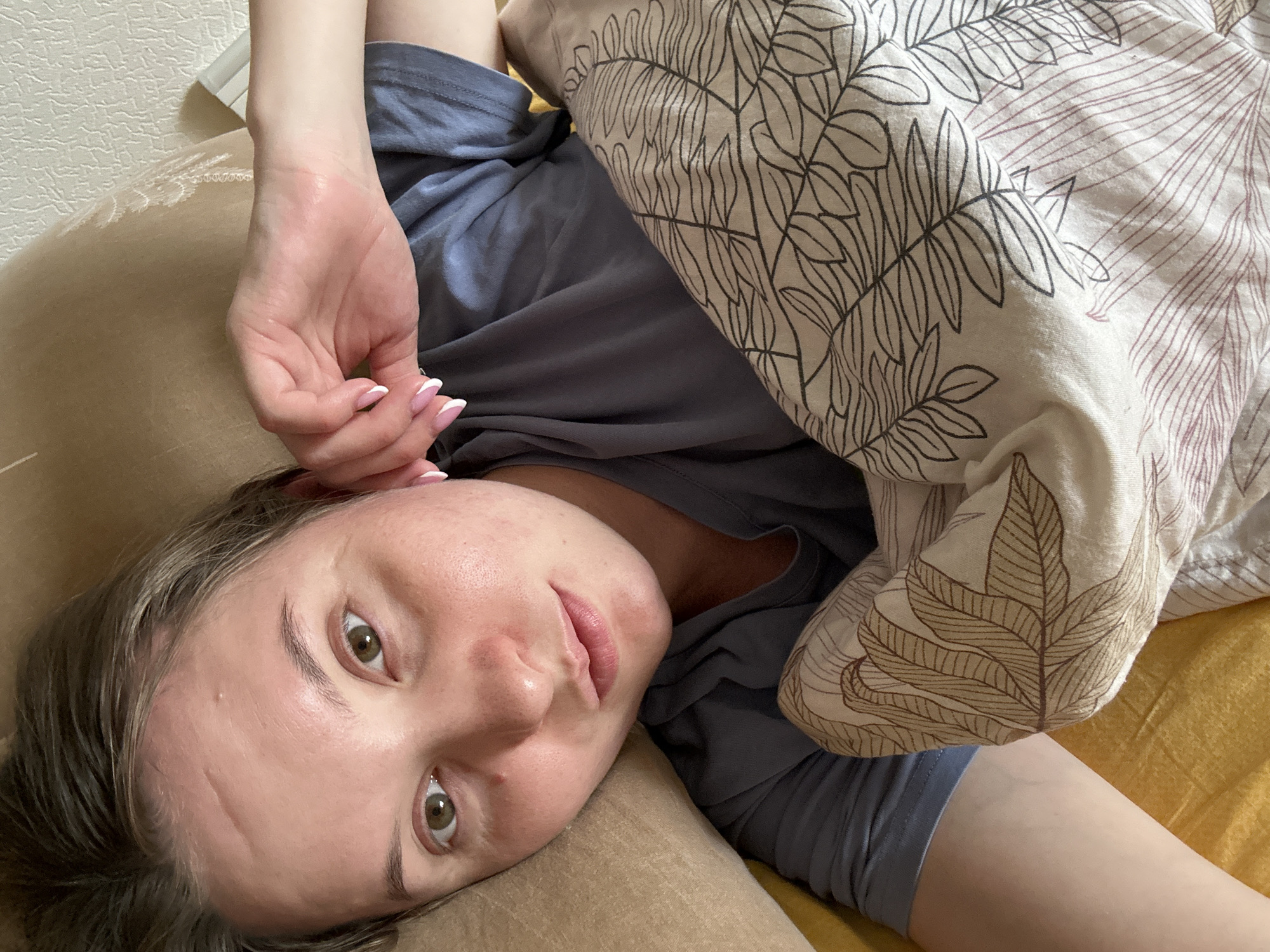 A woman is lying in bed, looking at the camera. She has blonde hair and green eyes, and she is wearing a grey t-shirt. She is covered with a blanket with a leaf pattern. The woman's left hand is resting on her chest, and her right arm is outstretched. The image appears to have been taken in a bedroom in Noviy, Russia.