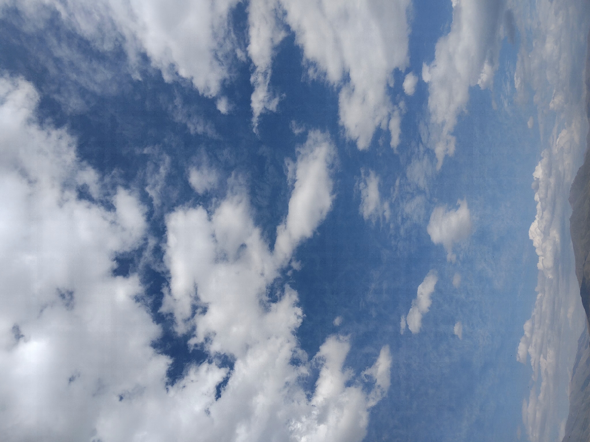 An image captures a wide expanse of blue sky filled with scattered white clouds, some dense and puffy, others thin and wispy. The lower portion of the image shows a distant, hazy mountain range with undulating peaks. The sky is the dominant element, with a clear gradient of blue. There are no people or man-made structures visible in the frame, suggesting an outdoor, natural landscape. The lighting indicates daylight, likely midday or afternoon, with the sun not directly visible but illuminating the clouds from above. The overall impression is one of a clear, cloud-filled sky over a mountainous region.