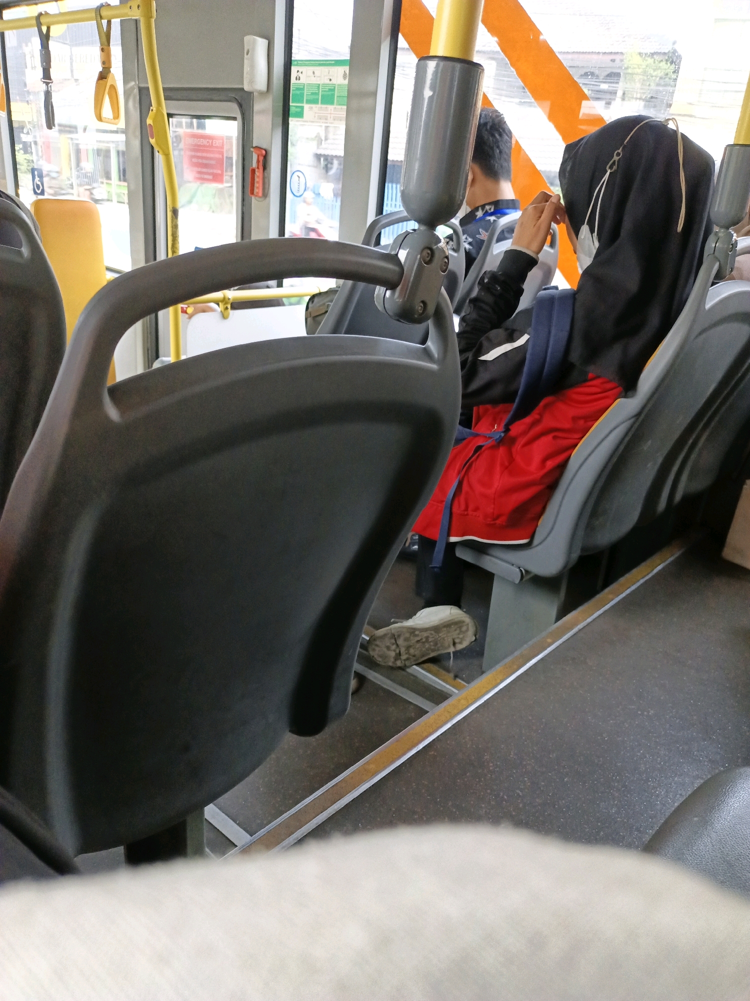 This image shows the interior of a public bus in South Tangerang, Indonesia. The focus is on the seating area, with the foreground dominated by the back of a dark gray bus seat. In the middle ground, a woman wearing a black hijab and a face mask is seated, with her right hand near her face. She is wearing a black jacket with white stripes and a bright red skirt. A dark blue backpack is visible behind her. To her left, a man is seated, wearing a black shirt with a white and blue pattern. He is facing away from the camera. The bus interior features yellow support poles and handrails, and large windows that offer a blurred view of the outside environment, which appears to be an urban setting with buildings and greenery. The floor of the bus is dark gray with yellow and silver strips along the aisle. A dirty white shoe is visible on the floor in front of the woman. The overall impression is a candid shot of passengers on public transportation in an Indonesian city.
